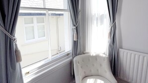 A corner room with a white armchair, a window with curtains, and a radiator. - Dukes Folly Hotel (Southport)