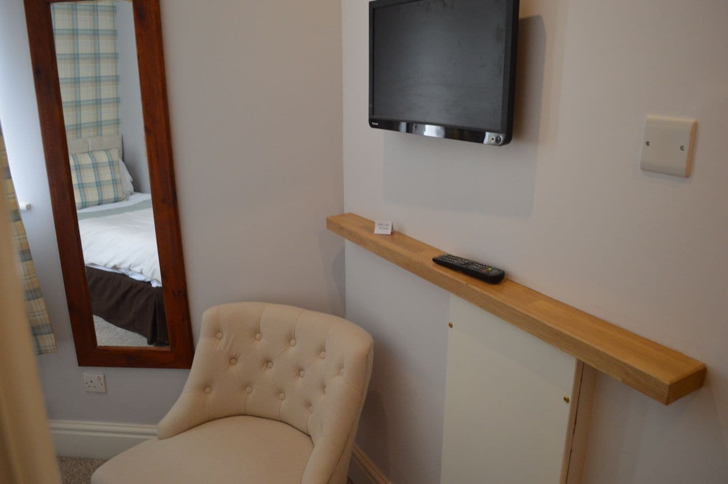 A hotel room with a bed, a chair, a television mounted on the wall, and a shelf with a remote control.