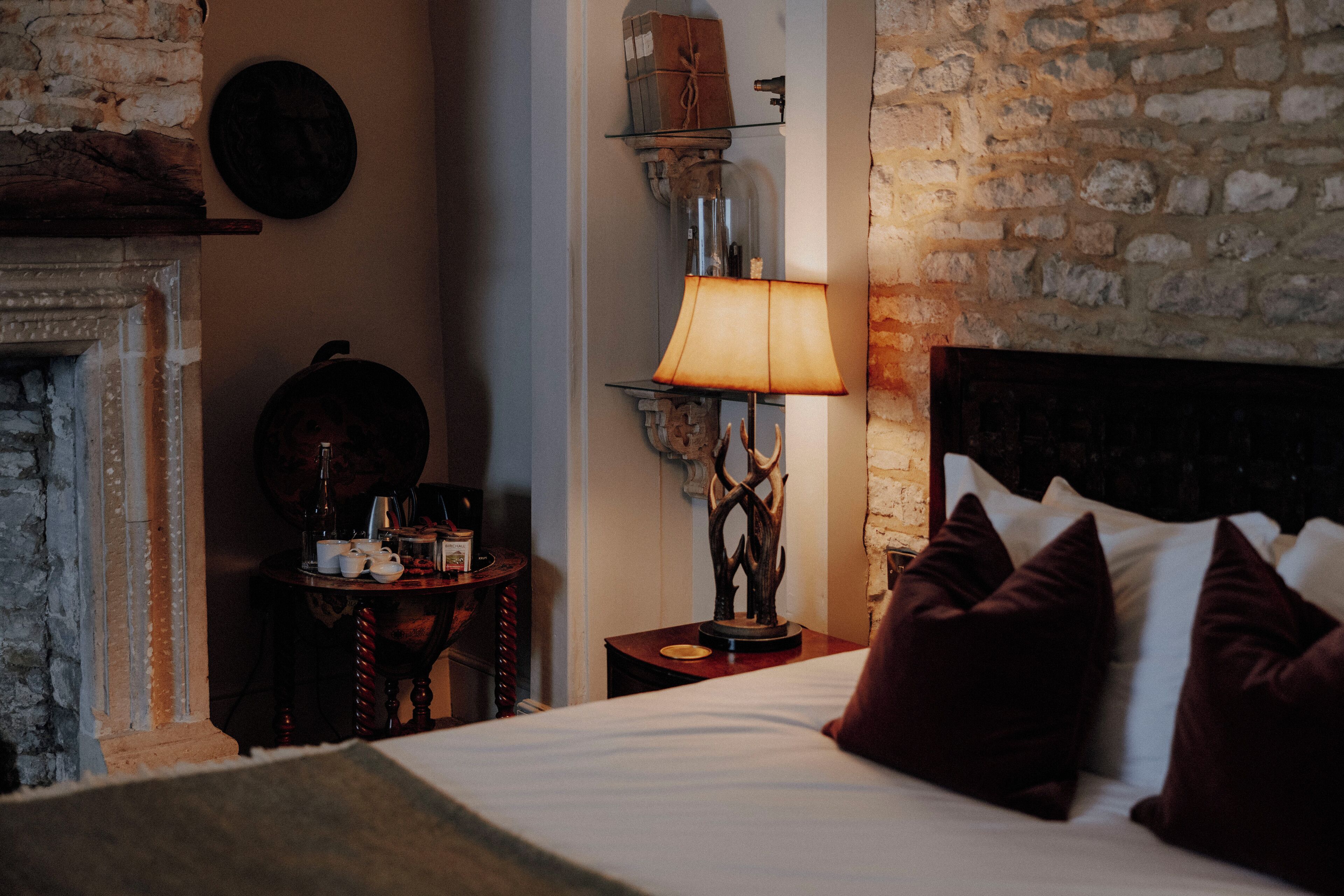 Un dormitorio con cama, almohadas, una lámpara, una mesita y una chimenea.