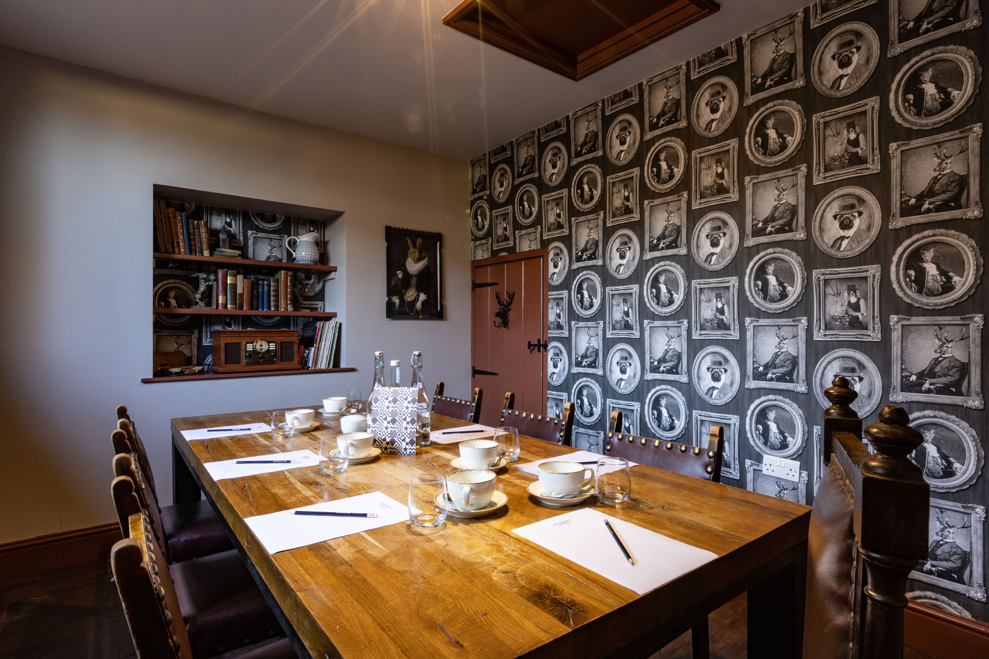 A dining room with a wooden table set for six, surrounded by chairs. A bookshelf and a wall of framed pictures are visible.