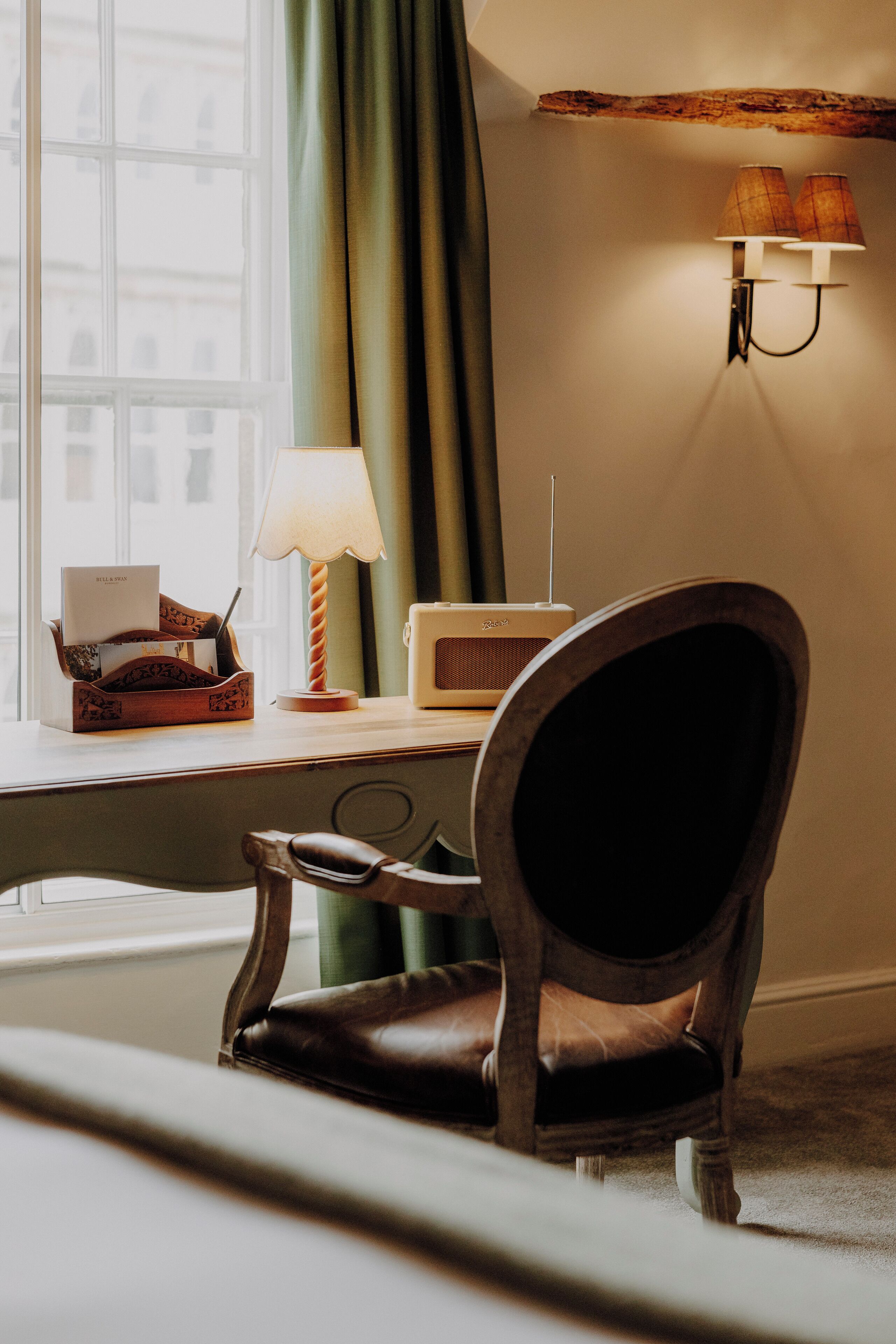 A room with a wooden chair, a desk with a lamp, a radio, and a window with curtains.
