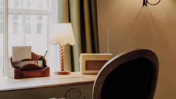 A room with a wooden chair, a desk with a lamp, a radio, and a window with curtains.