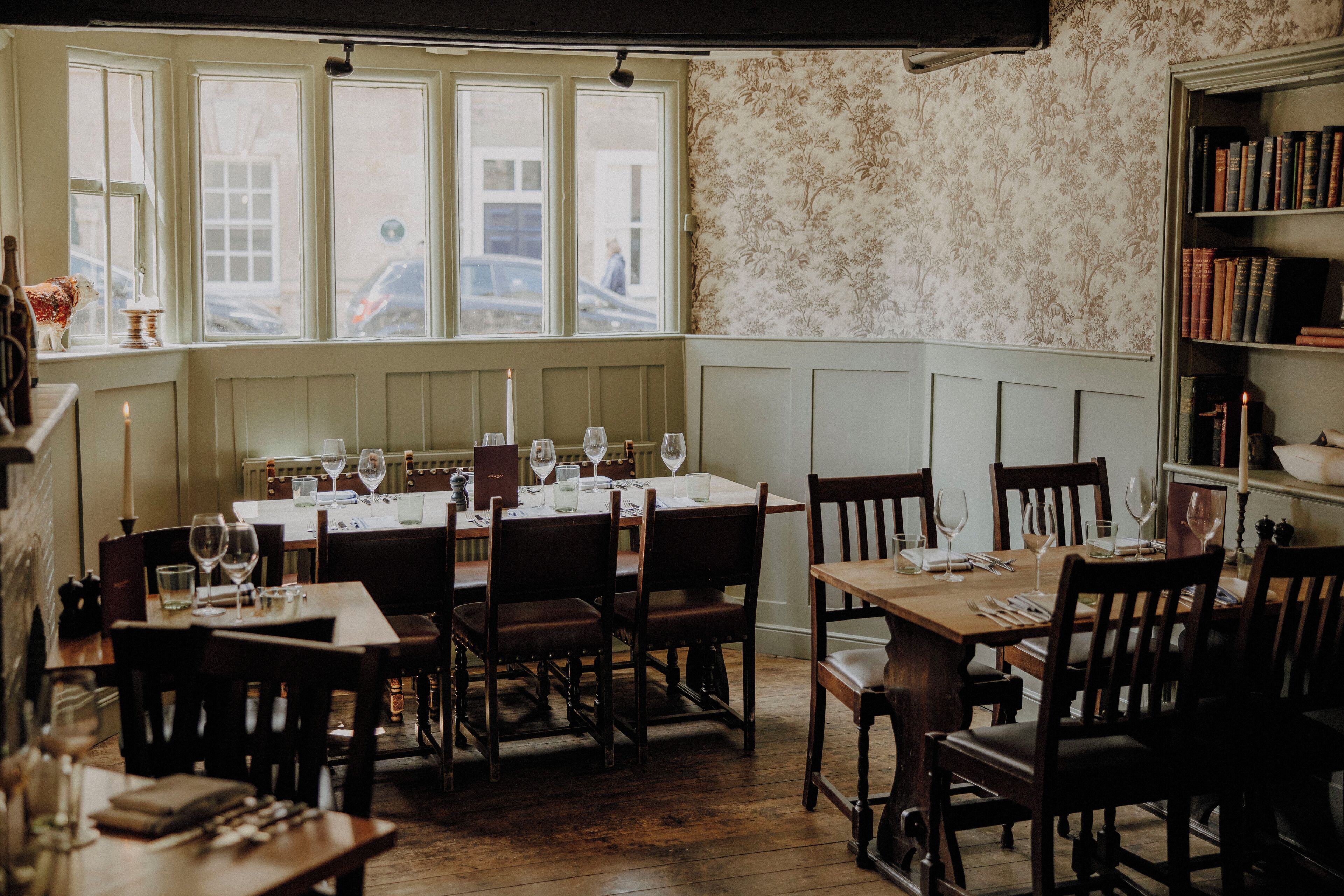 A dining area with wooden tables and chairs, a bookshelf, and a view through the window.