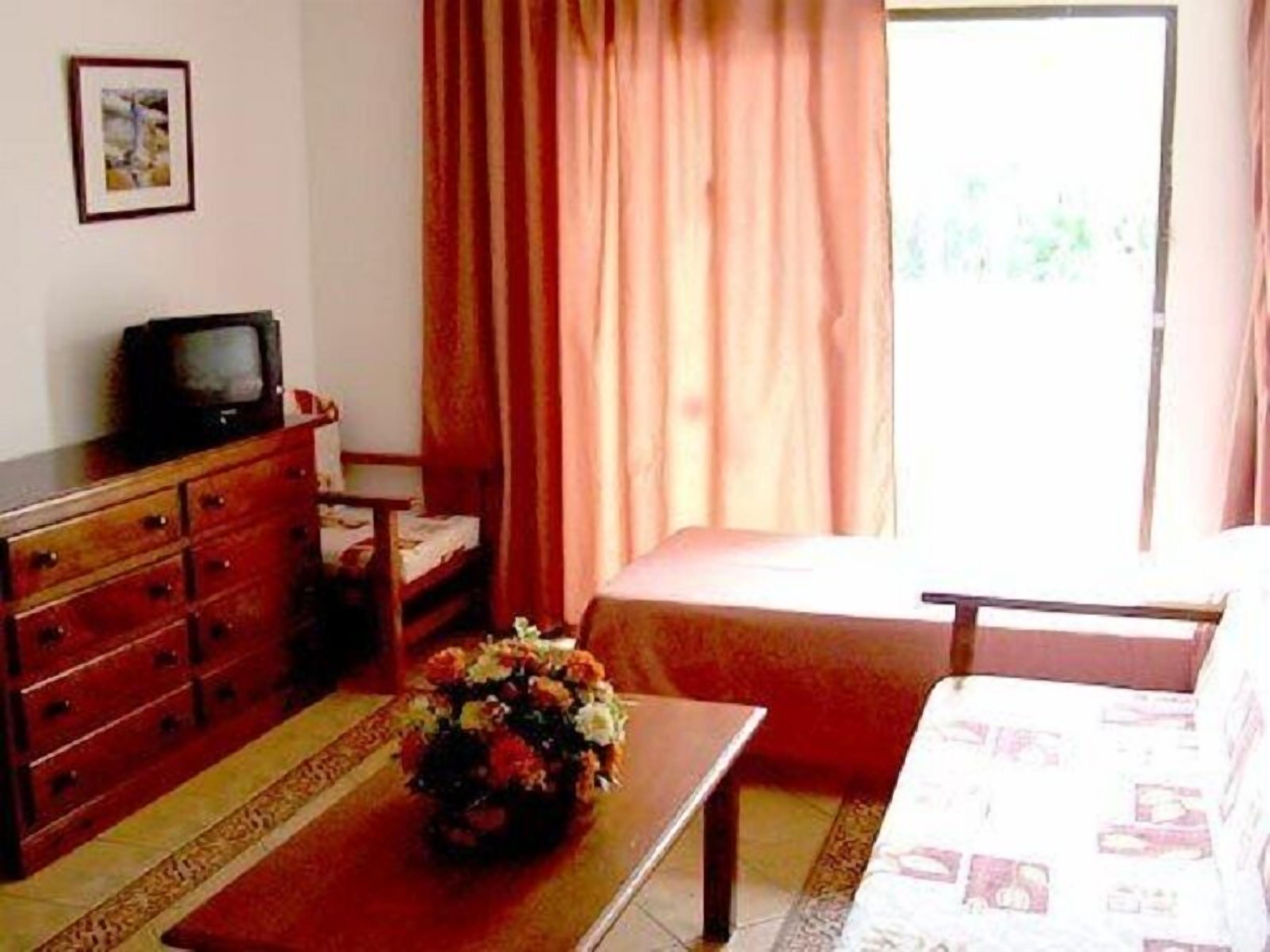 Una habitación con un aparador de madera, un televisor encima, un florero sobre una mesa, un sofá y una ventana con cortinas.