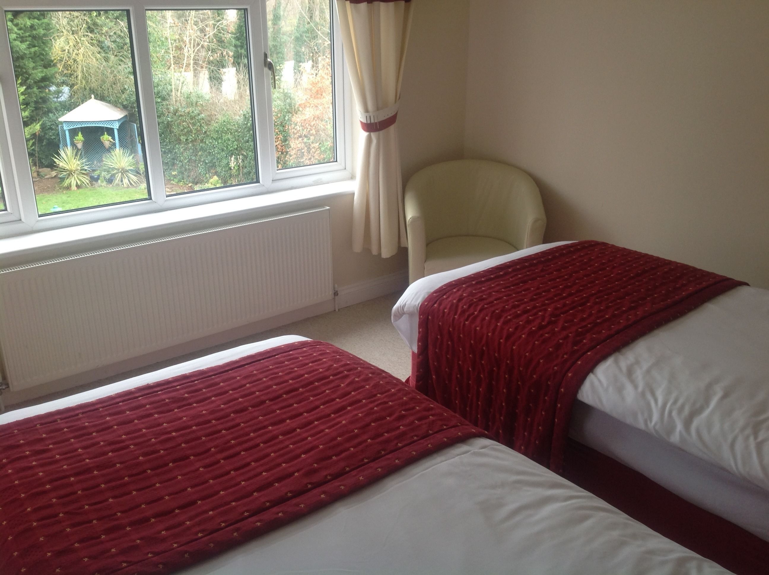 A bedroom with two beds, a chair, a window with curtains, and a view of greenery outside.