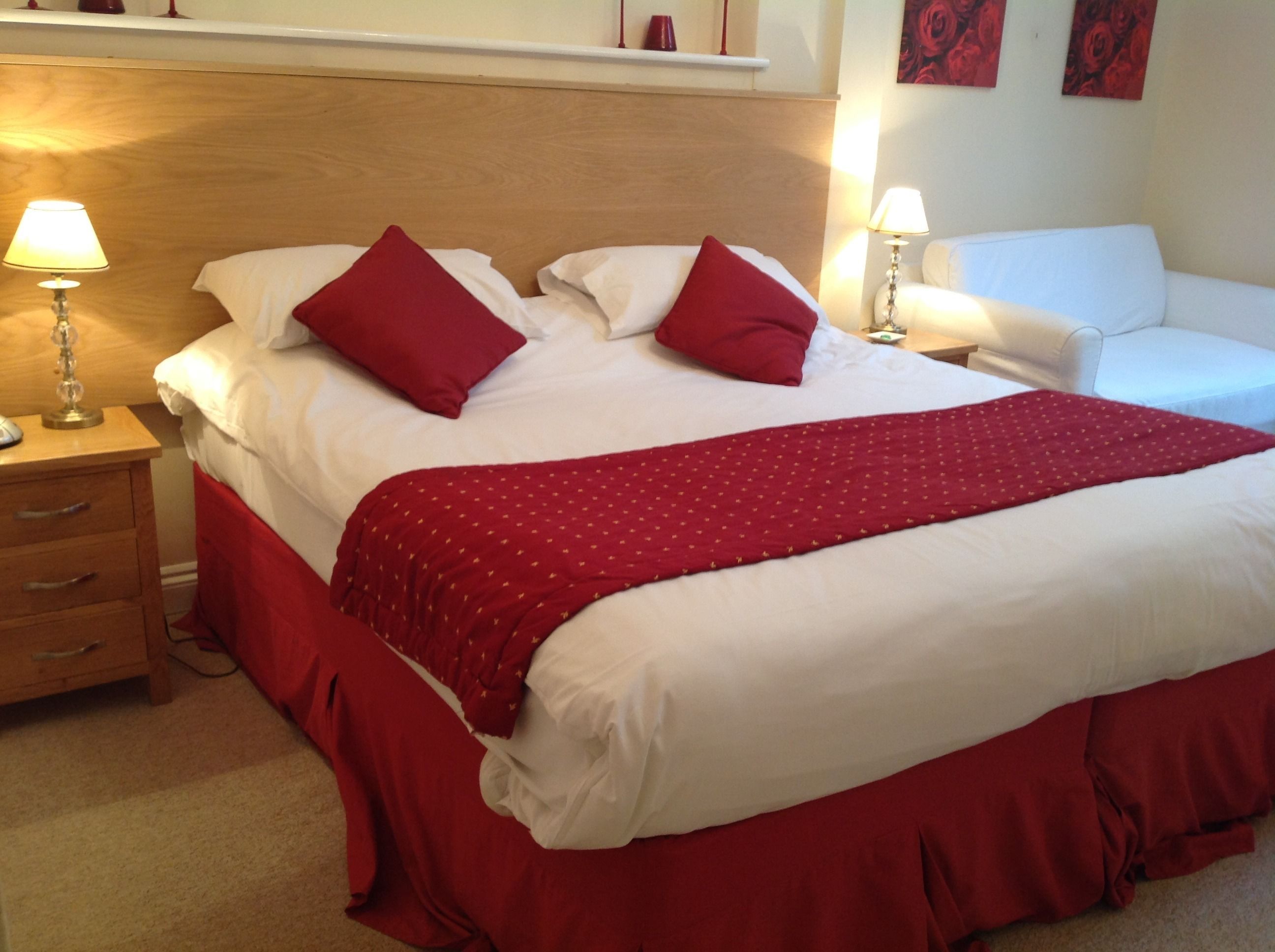 A neatly made bed with red bedding and pillows, a wooden headboard, a bedside table with a lamp, and a white armchair.