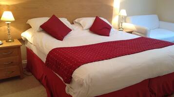 A neatly made bed with red bedding and pillows, a wooden headboard, a bedside table with a lamp, and a white armchair.