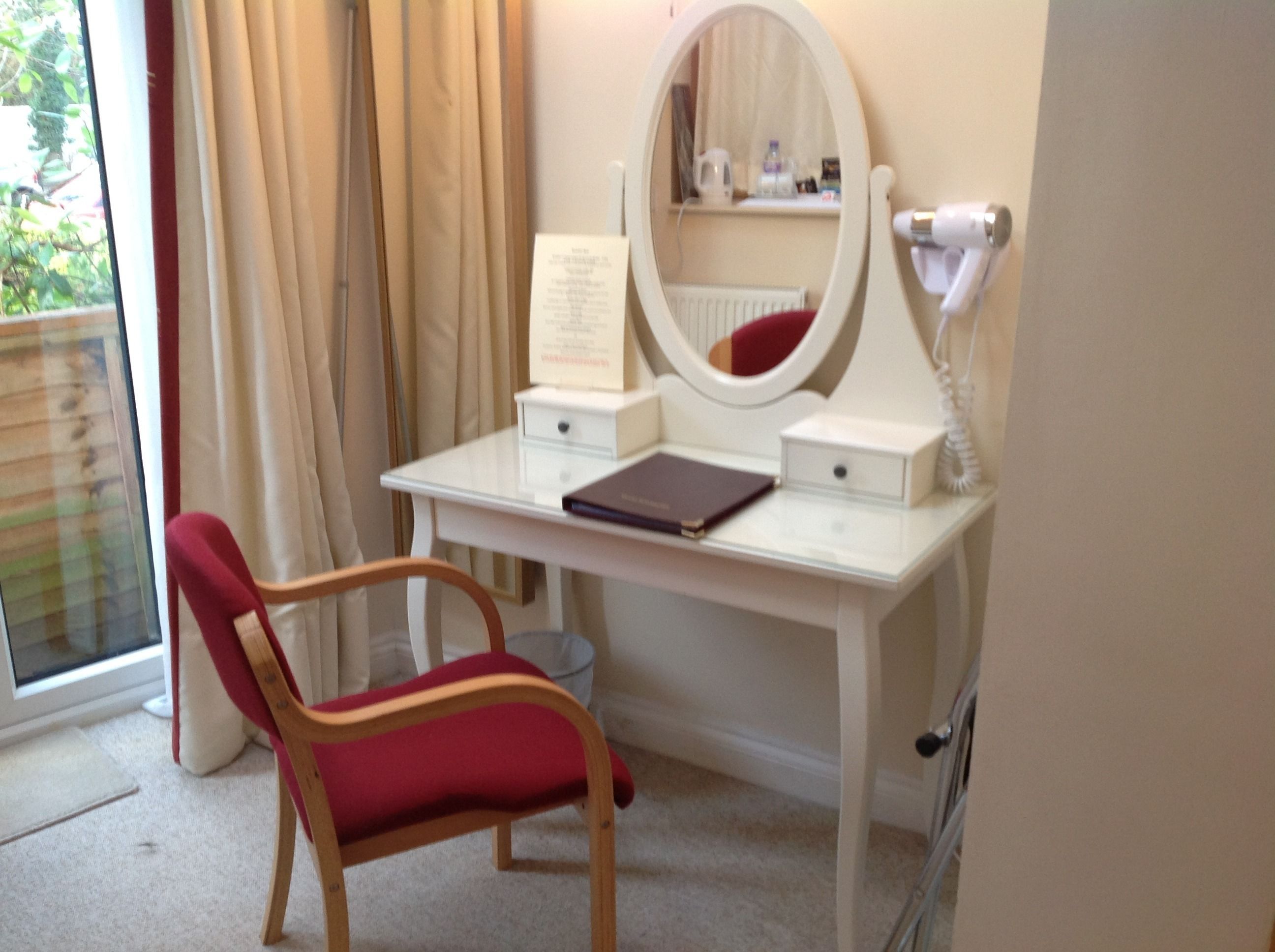 A room with a white vanity table, a red chair, a mirror, and a hairdryer.