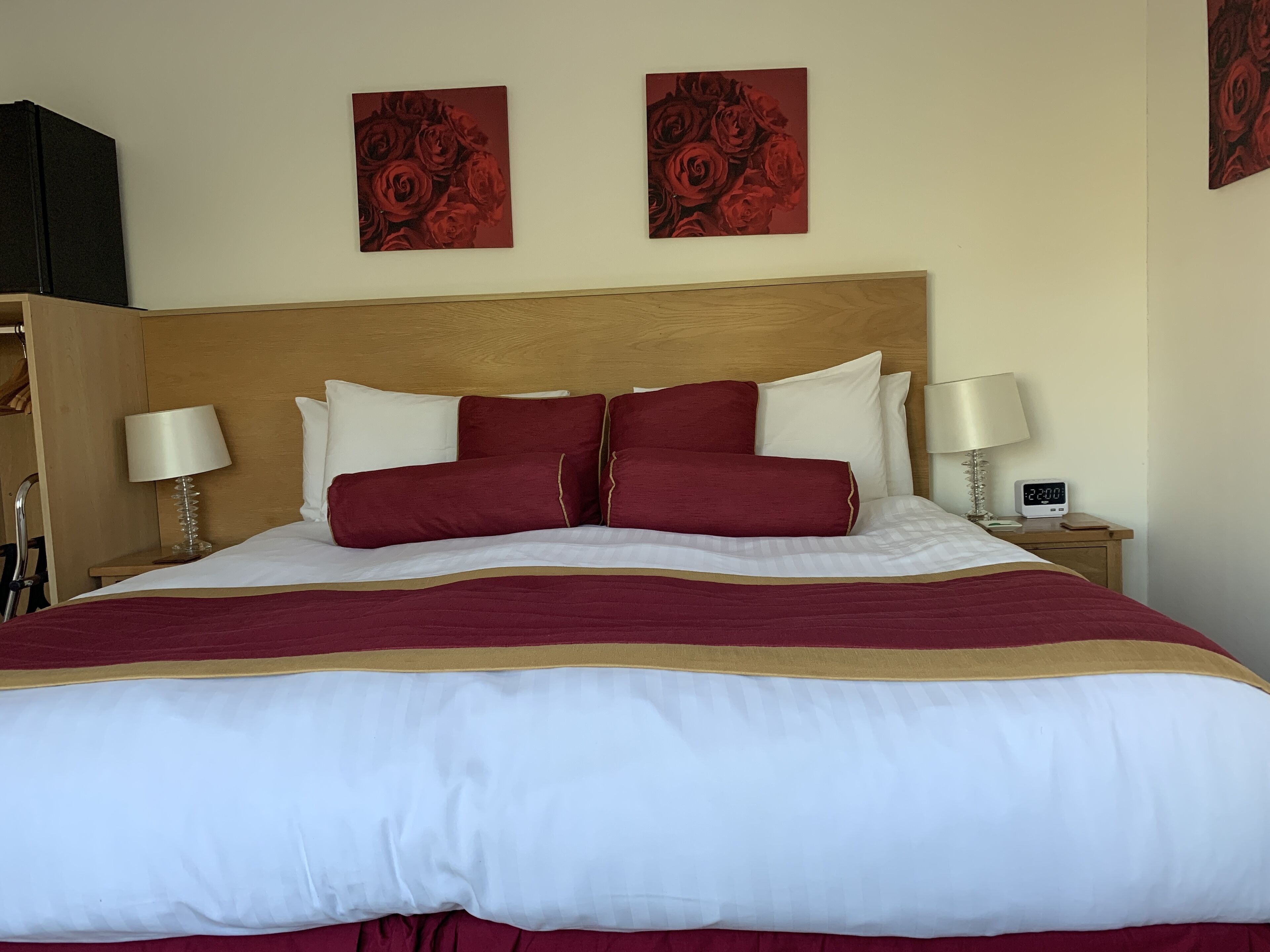 Un lit bien fait, avec une tête de lit en bois, une literie blanche et bordeaux, et deux tableaux décoratifs aux motifs floraux rouges accrochés au-dessus.