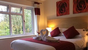 Habitación de hotel con una cama grande, almohadas rojas, una manta roja y dos toallas con forma de cisne. Se ve una ventana con cortinas, una lámpara y un cuadro de una rosa en la pared.