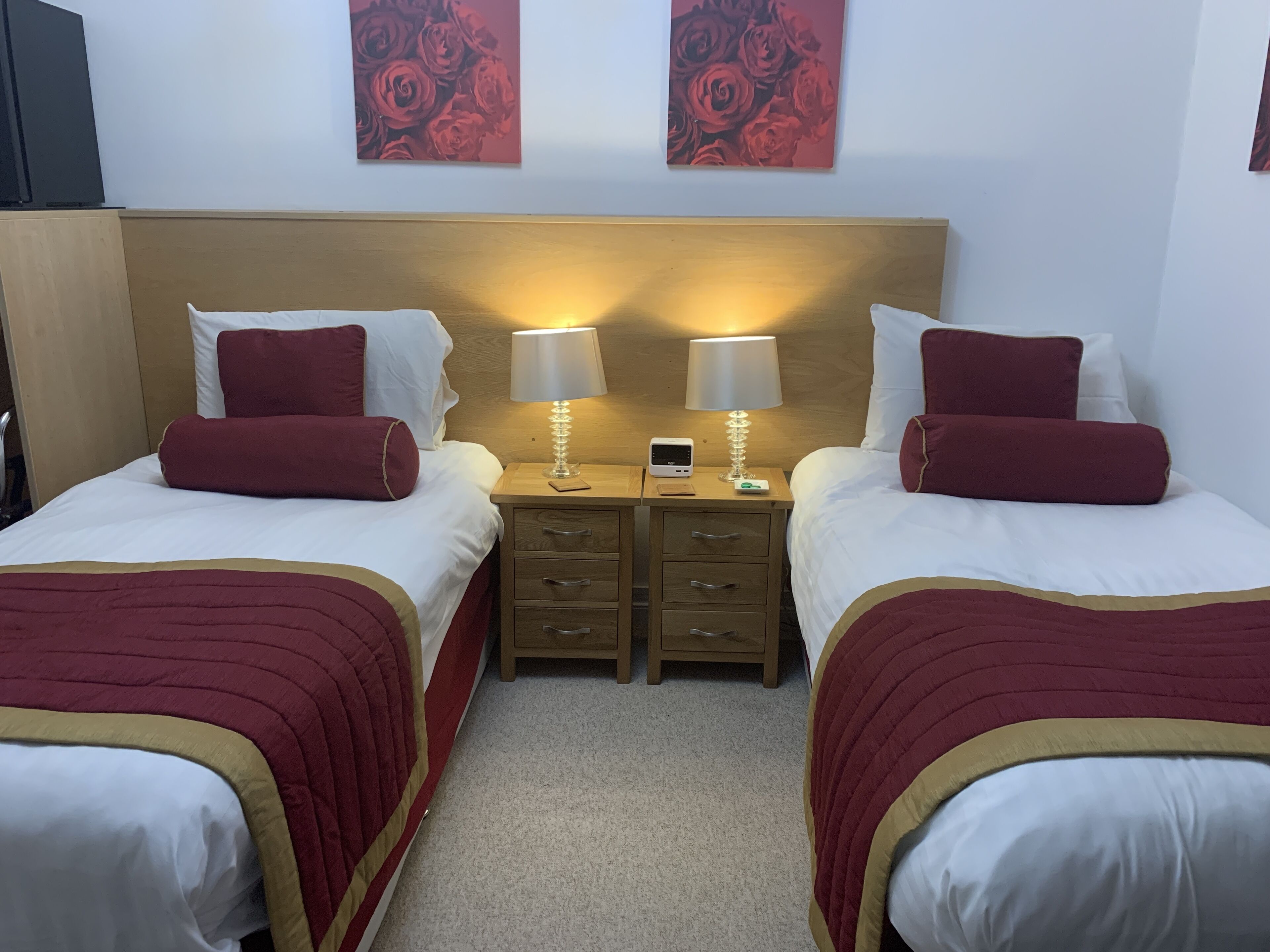 A hotel room with two beds, each with a red and gold bedspread, white pillows, and a wooden headboard. There are two bedside tables with lamps, a small clock, and a digital device. The walls are adorned with red rose paintings.