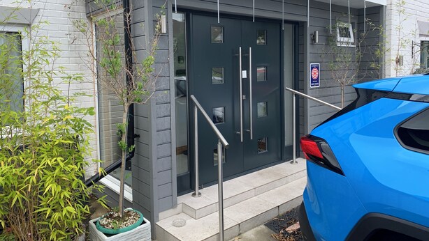 Un edificio moderno con una puerta gris oscuro, un coche azul estacionado adelante y un pequeño jardín con plantas de bambú.