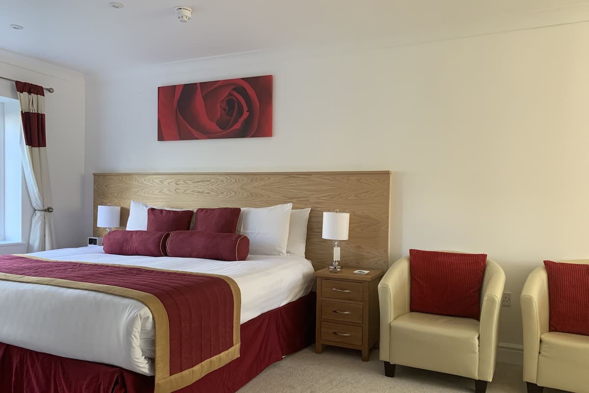 A hotel room with a large bed, two bedside tables with lamps, a wooden headboard, and two armchairs.
