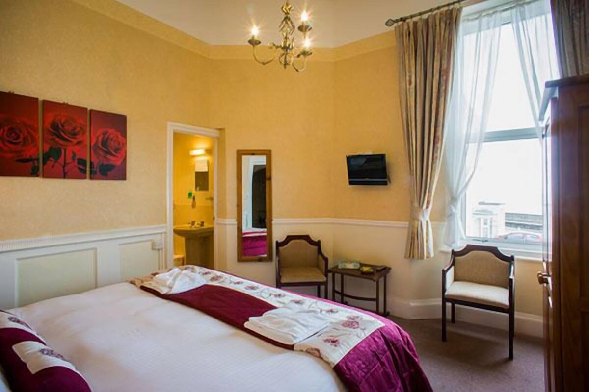 A hotel room with a large bed, a chandelier, a mirror, two chairs, and a window with curtains.
