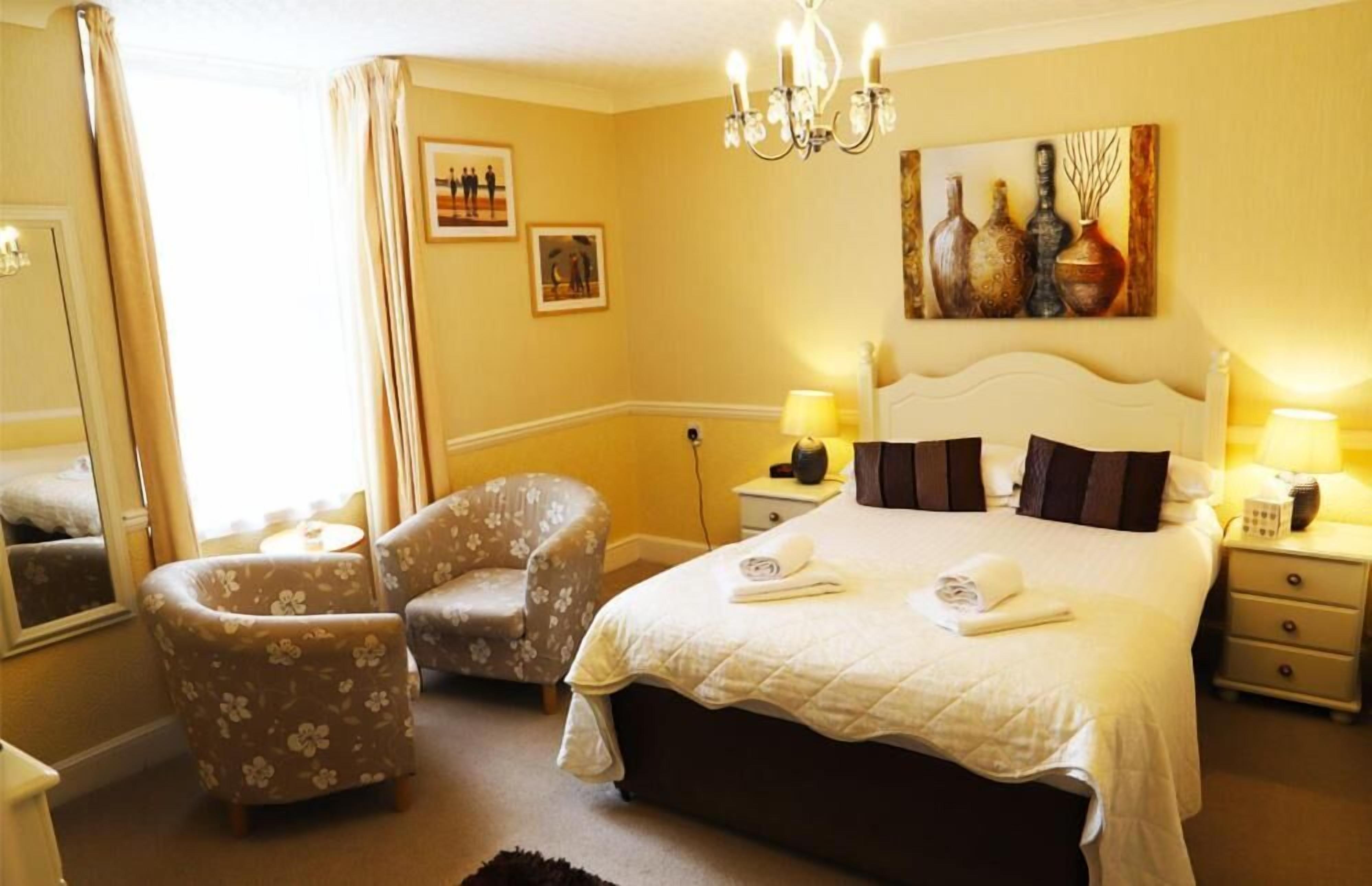 A hotel room with a large bed, two armchairs, a chandelier, bedside lamps, and artwork on the walls.