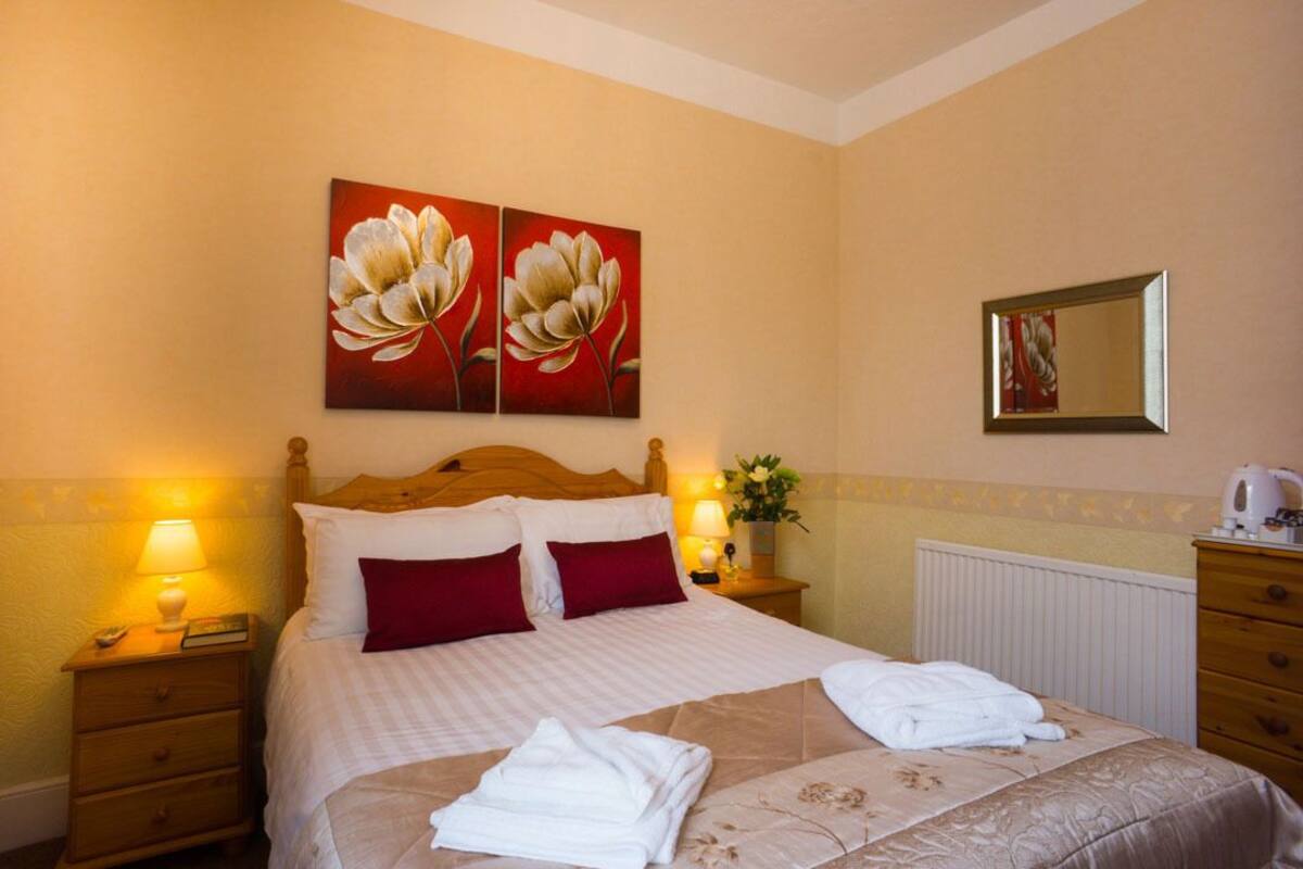 A hotel room with a bed, two pillows, a nightstand with a lamp, a dresser, and a mirror.