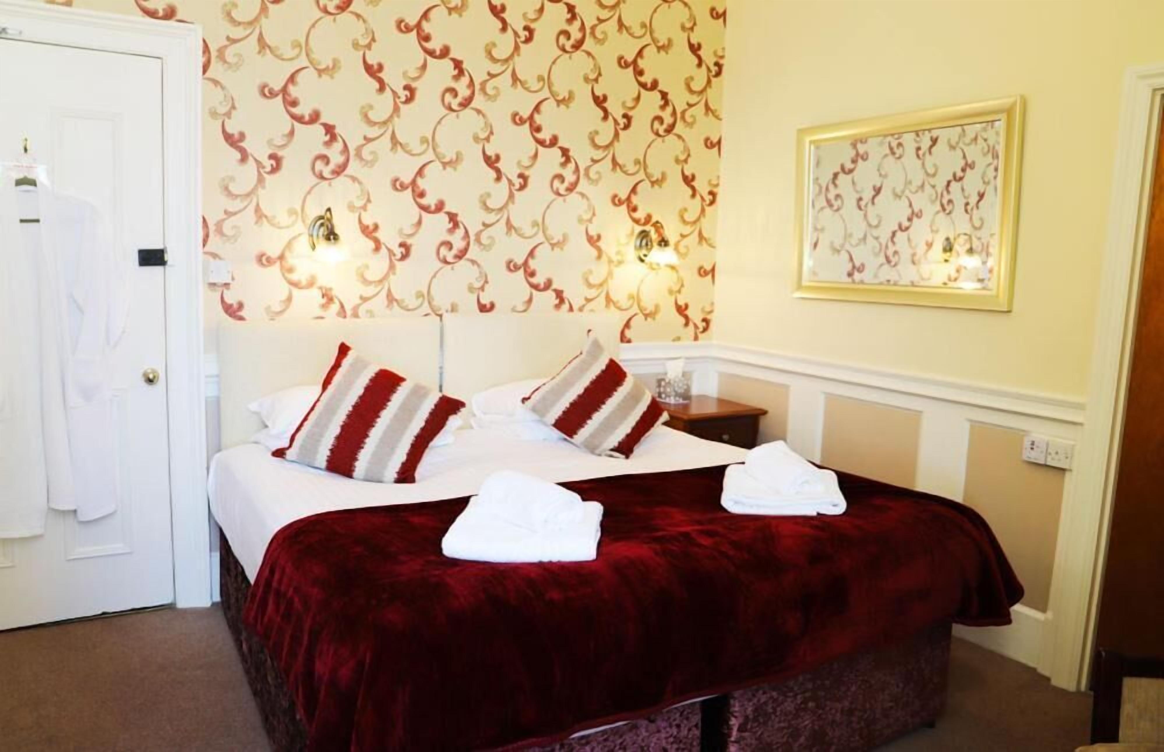 A hotel room with a large bed, two pillows, a red blanket, a white towel, a nightstand, a wall clock, and a framed picture.