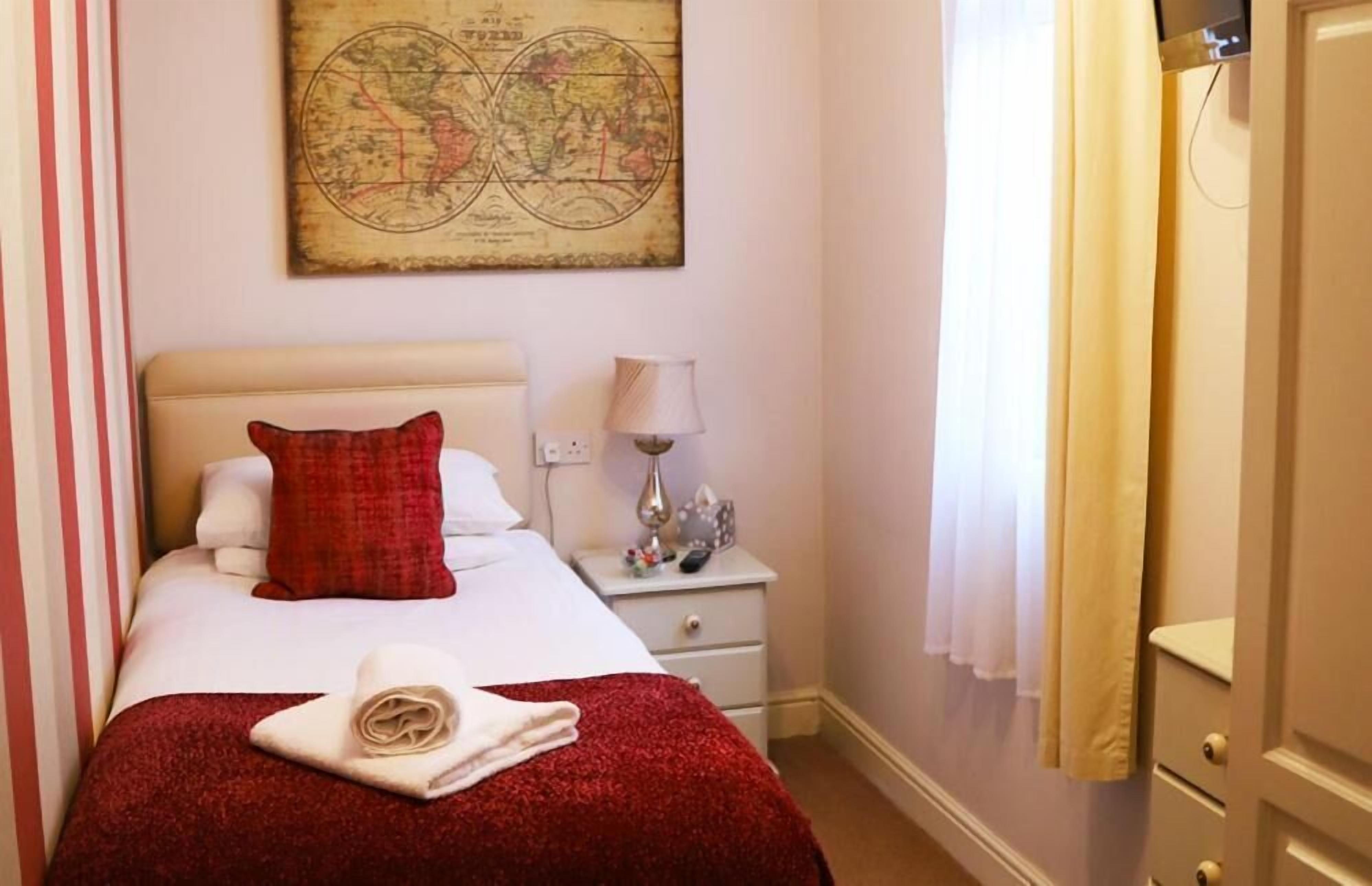 A neatly made bed with a red blanket and pillows, a bedside lamp, a small table with a lamp, and a world map on the wall.
