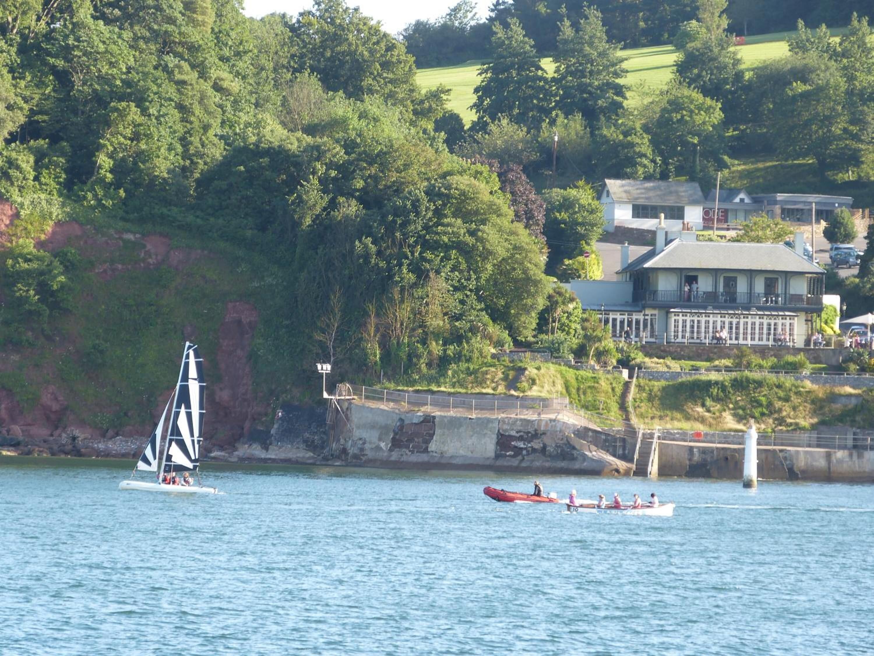 A coastal scene with a sailboat and a rowboat on the water, a rocky shoreline, and a building with a balcony overlooking the water.