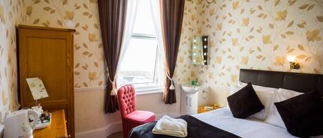 A hotel room with a bed, a vanity with a mirror, a red chair, a wooden cabinet with a kettle, and a window with curtains.