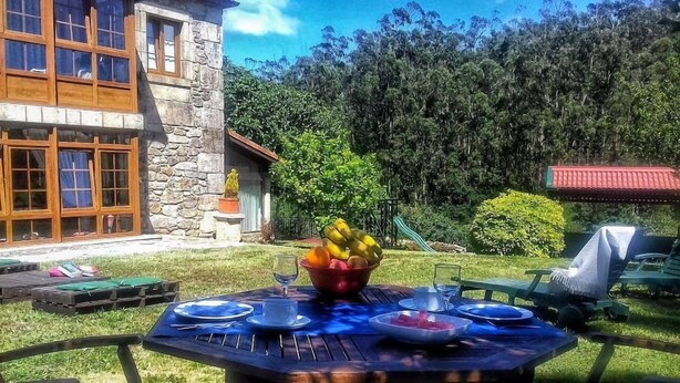 An outdoor dining area with a table set for four, surrounded by chairs, in a garden with a stone building in the background.