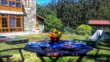 Un área de comedor al aire libre con una mesa preparada para cuatro personas, rodeada de sillas, en un jardín con un edificio de piedra al fondo.