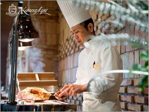 A chef in a white uniform with a badge, preparing food in a kitchen with brick walls and a metal stove. - Hotel Kawakyu (Shirahama)