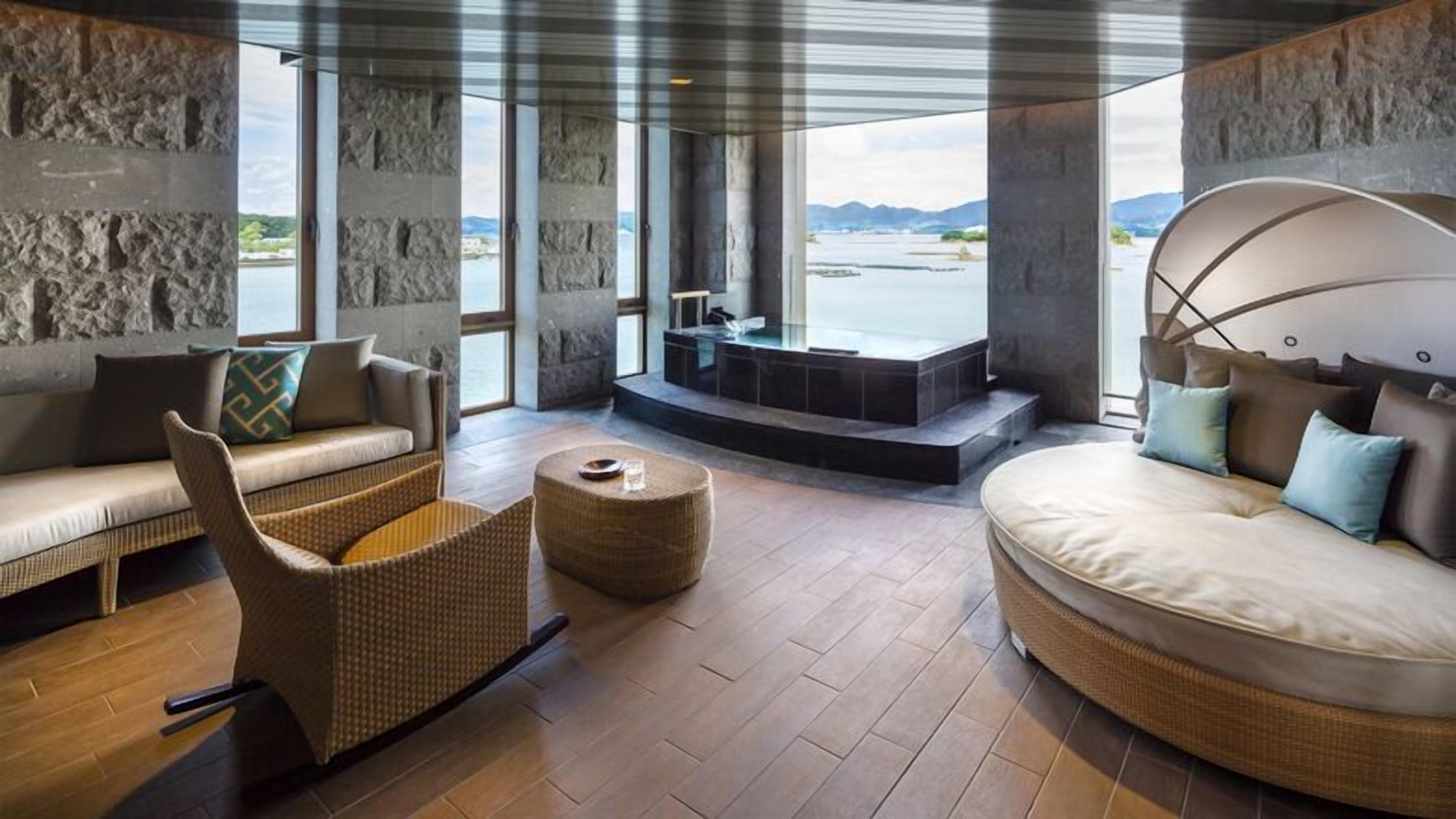 Une chambre moderne avec un mur en pierre, un canapé, un fauteuil, un lit rond et une vue sur un plan d’eau.