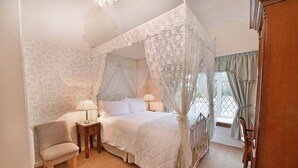 A bedroom with a four-poster bed, a bedside table with a lamp, a chair, and a window with curtains. - Virginia Lodge (Stratford-upon-Avon)