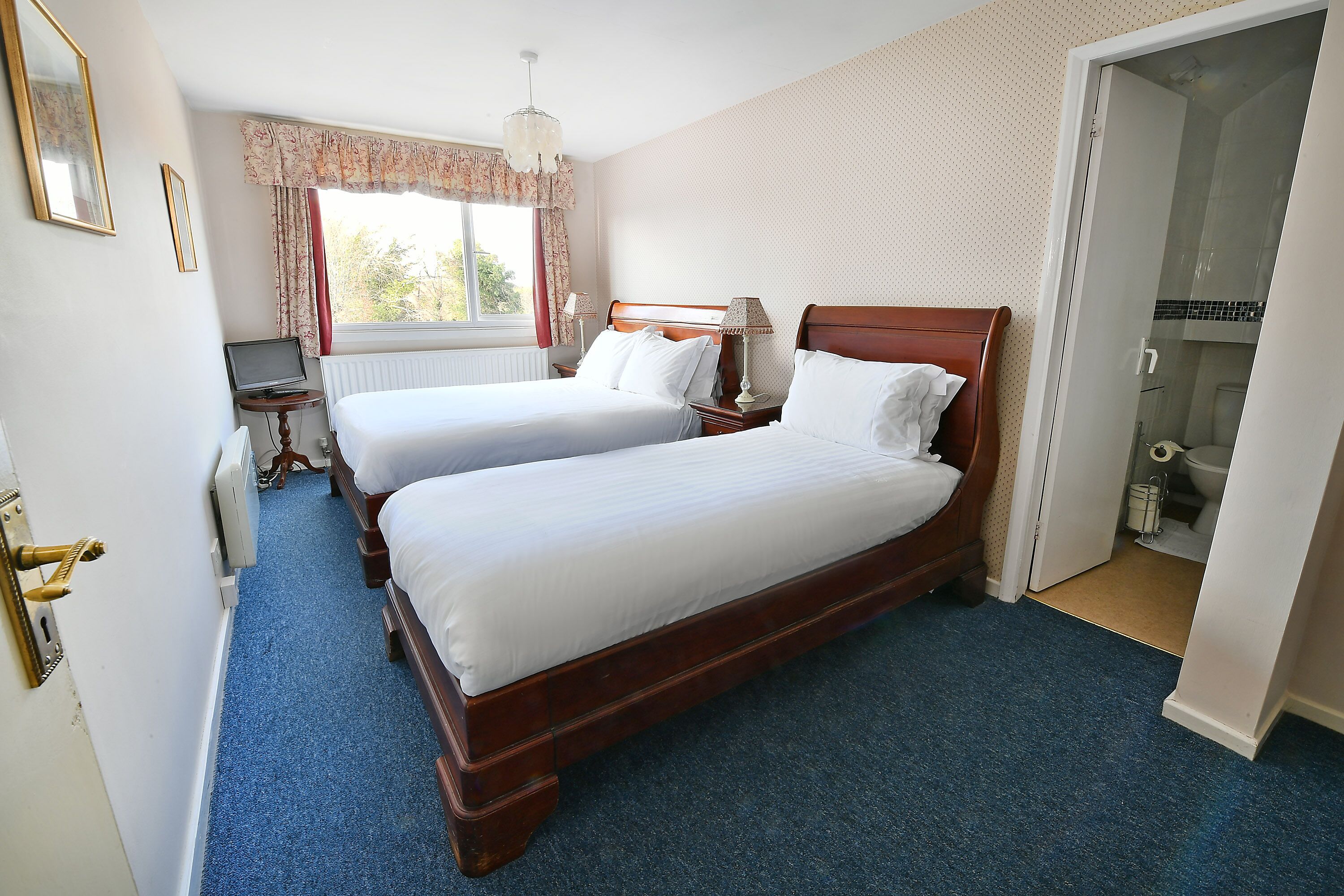 A hotel room with two single beds, a desk with a laptop, a window with curtains, and a shower area.