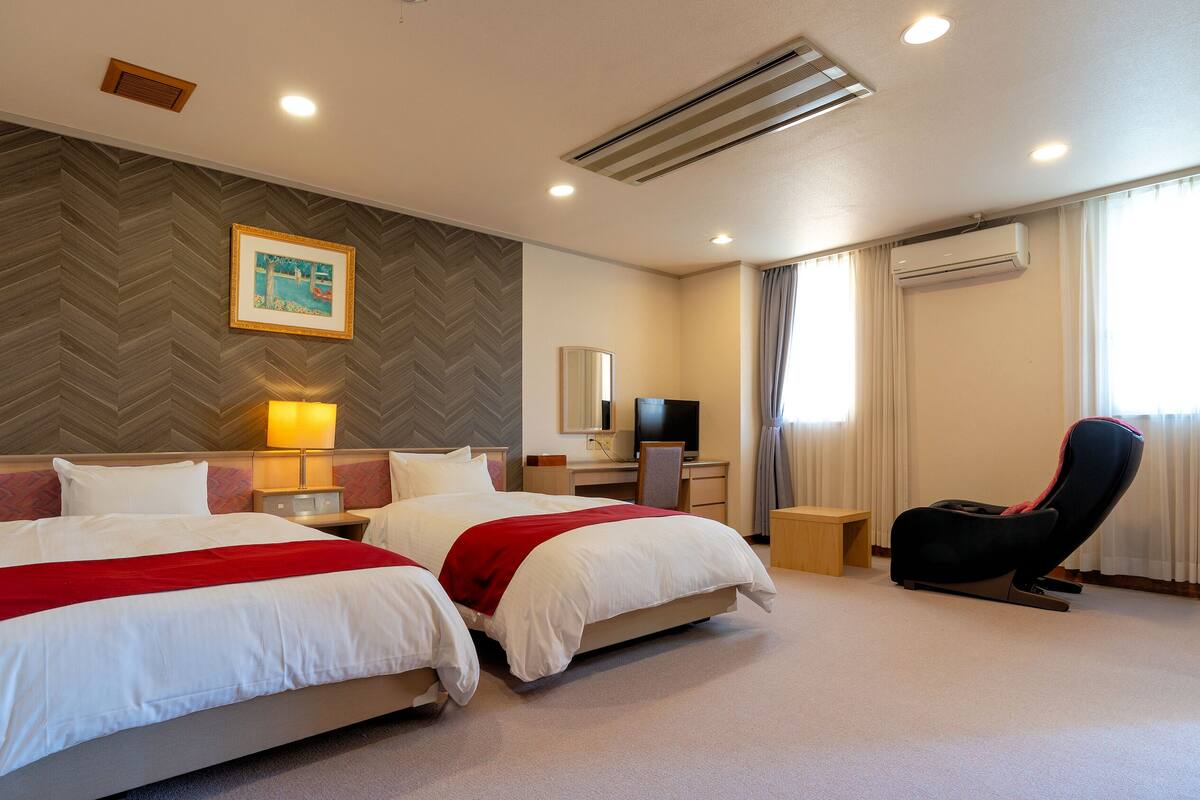 A hotel room with two beds, a desk, a chair, a TV, and an air conditioning unit.