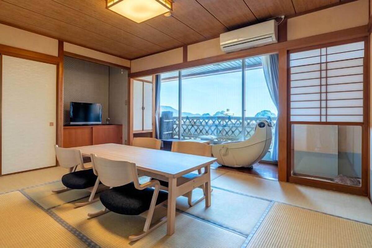 A traditional Japanese-style room with tatami flooring, a low table with chairs, a television, and a balcony with a view.