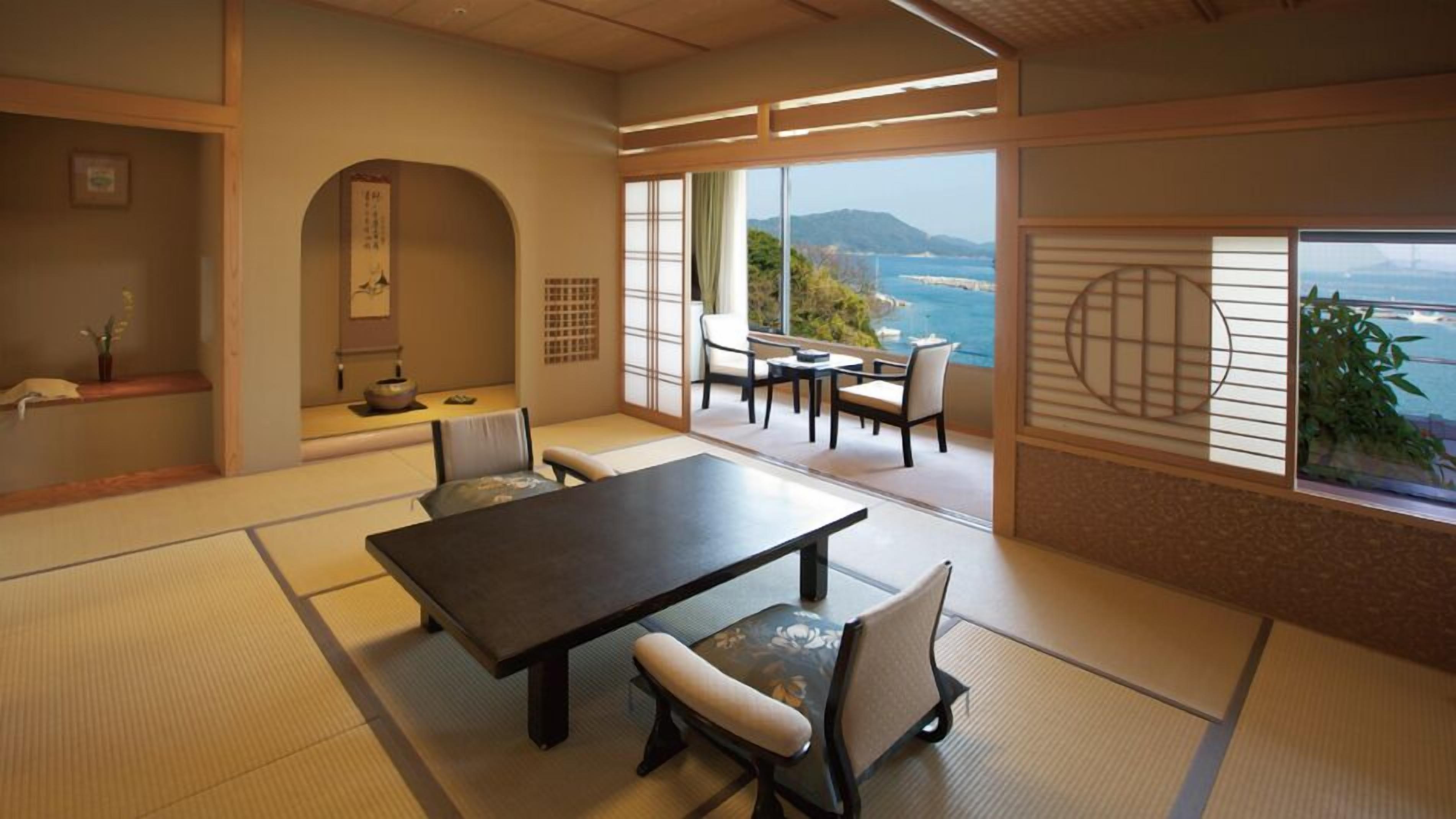 A traditional Japanese room with tatami flooring, a low table with chairs, a sliding door with a view, and a small alcove with a shelf and decorative items.