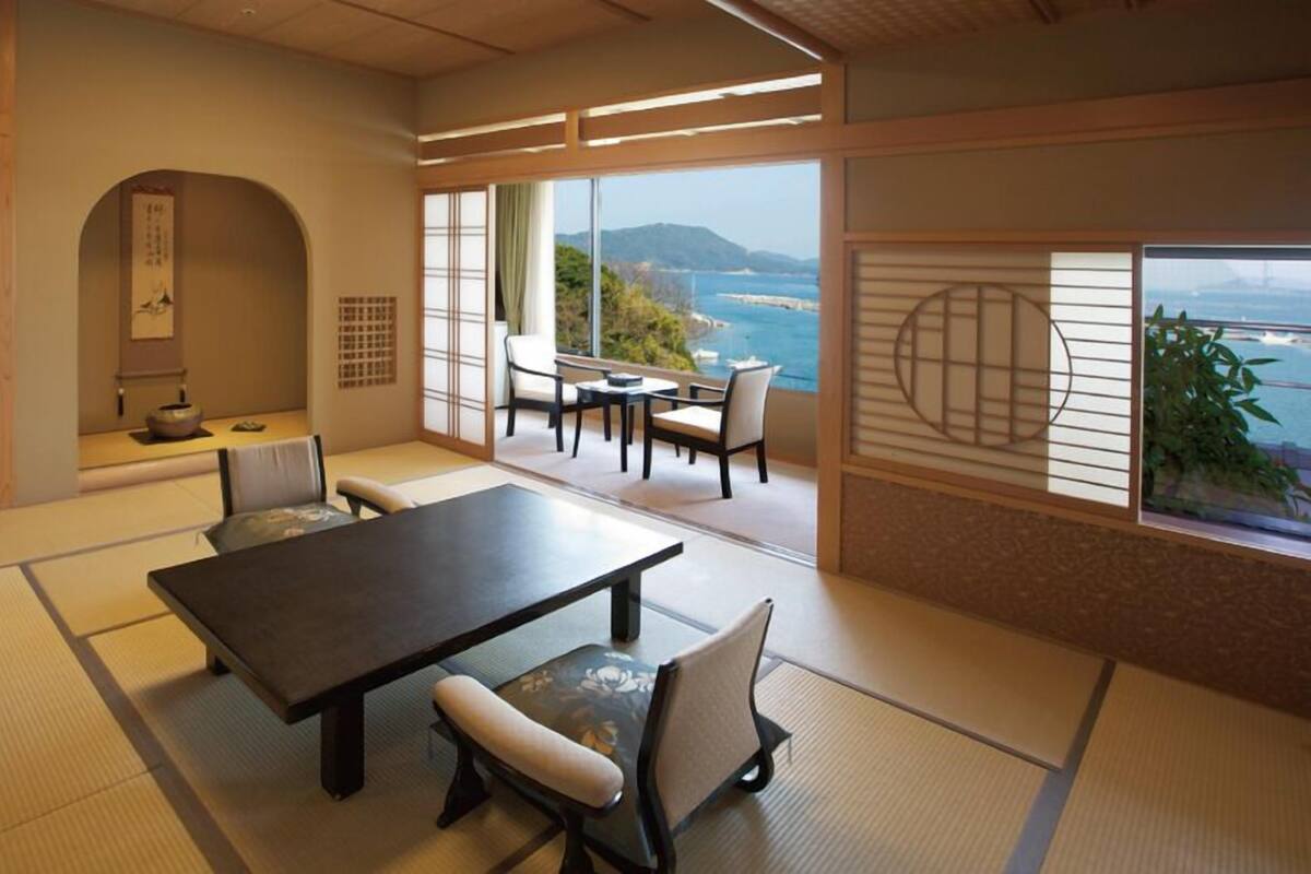 A traditional Japanese room with tatami flooring, a low table with chairs, a sliding door with a view, and a small alcove with a shelf and decorative items.