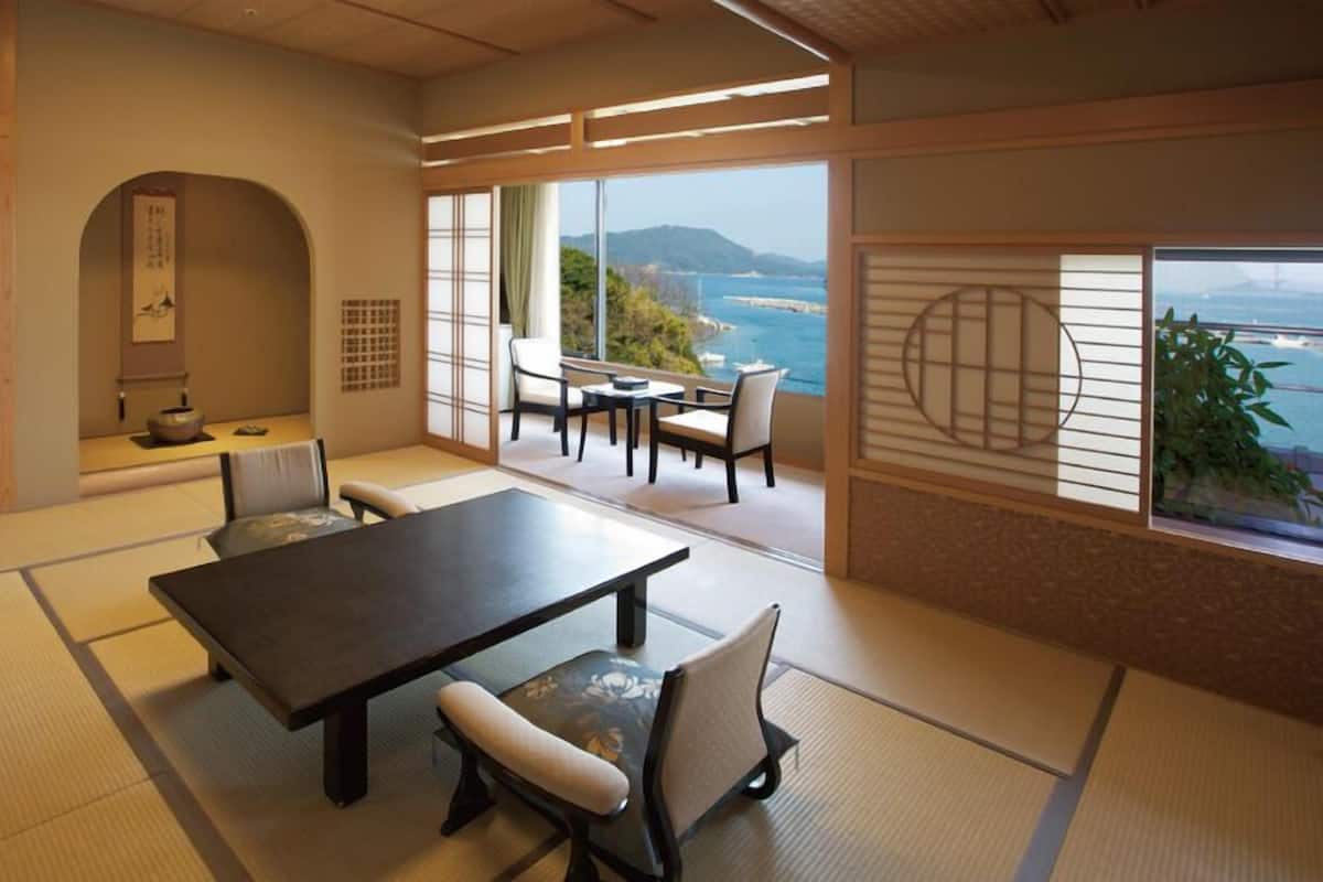 Una habitación tradicional japonesa con suelo de tatami, una mesa baja con sillas, una puerta corrediza con vistas, y un pequeño rincón con un estante y objetos decorativos.