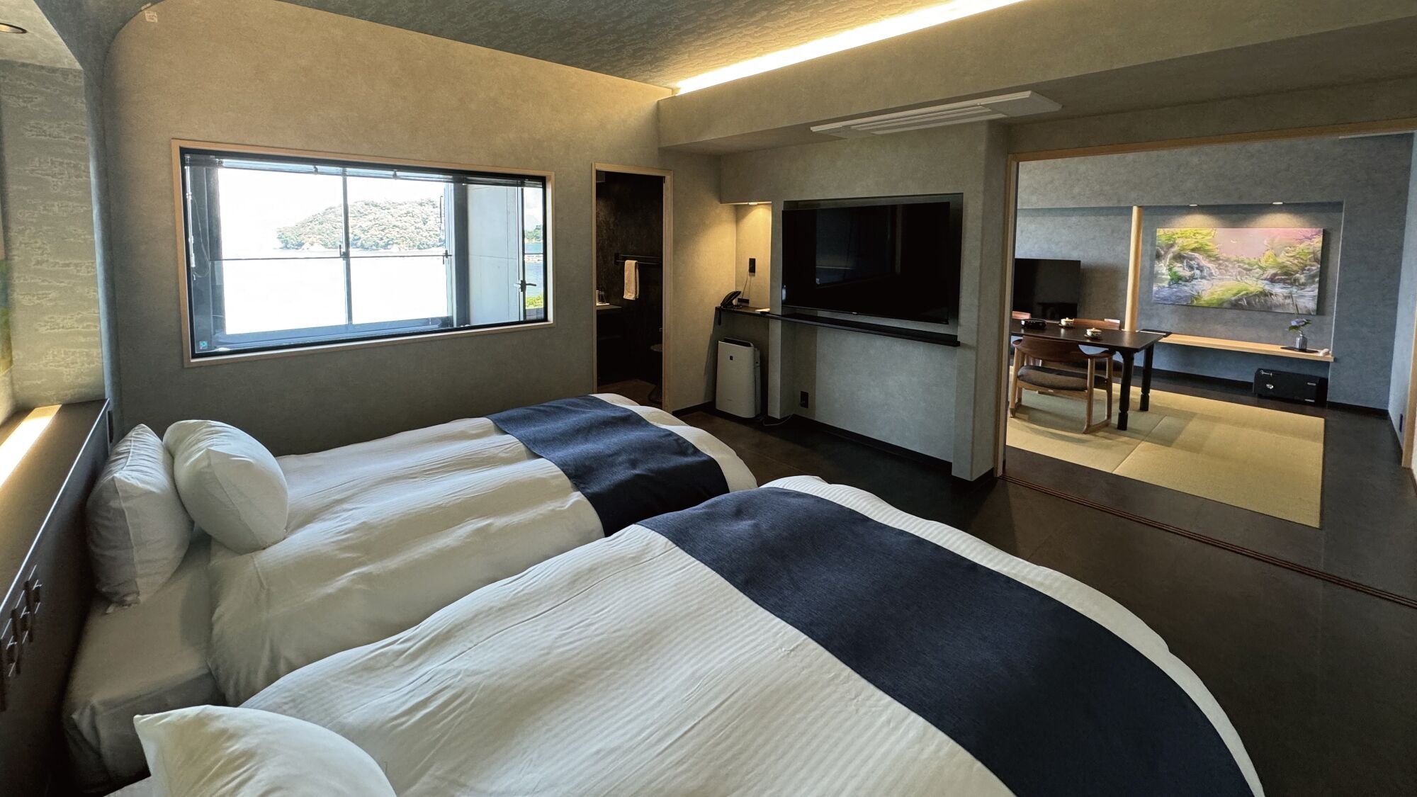 A modern hotel room with two beds, a kitchenette, a TV, and a large window.