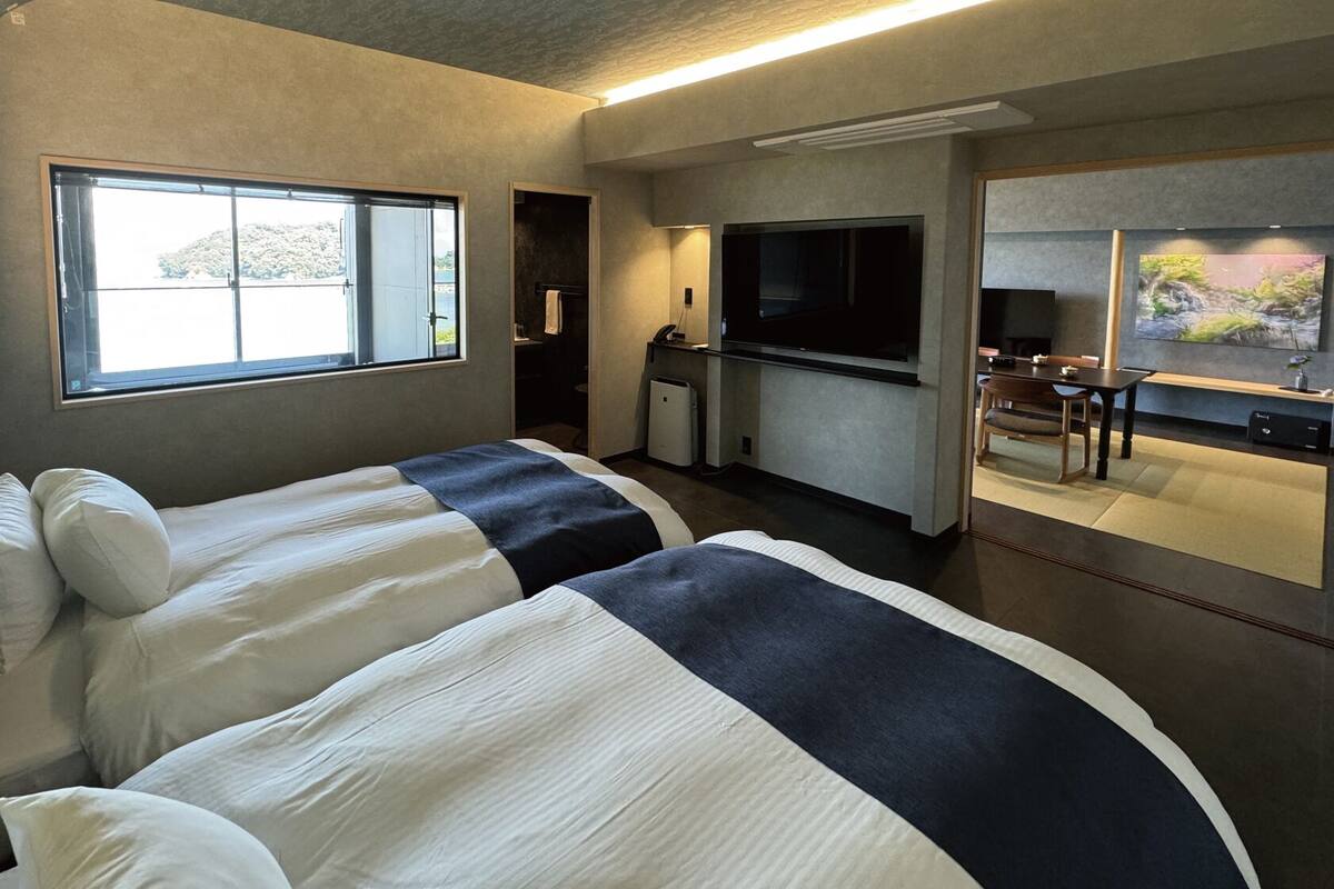 A modern hotel room with two beds, a kitchenette, a TV, and a large window.