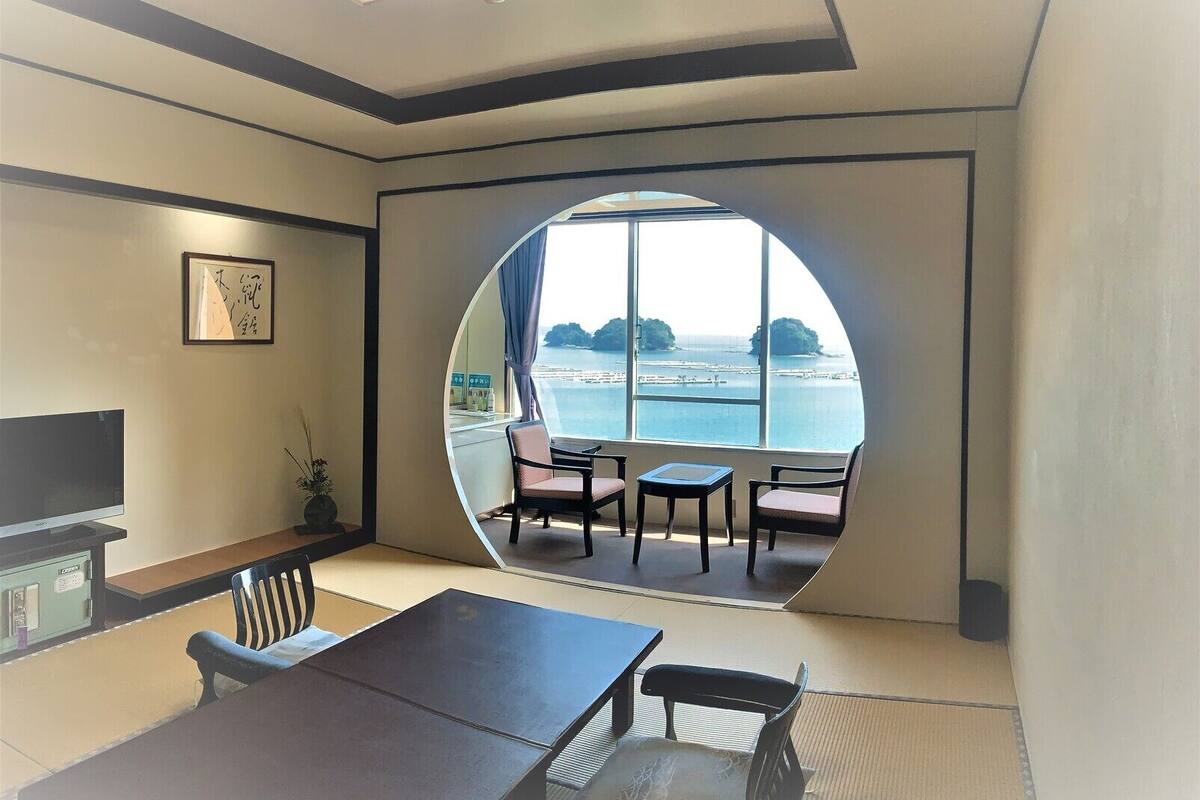 A room with a large circular window offering a view of the sea, a flat-screen TV, a dining table, and a sofa.