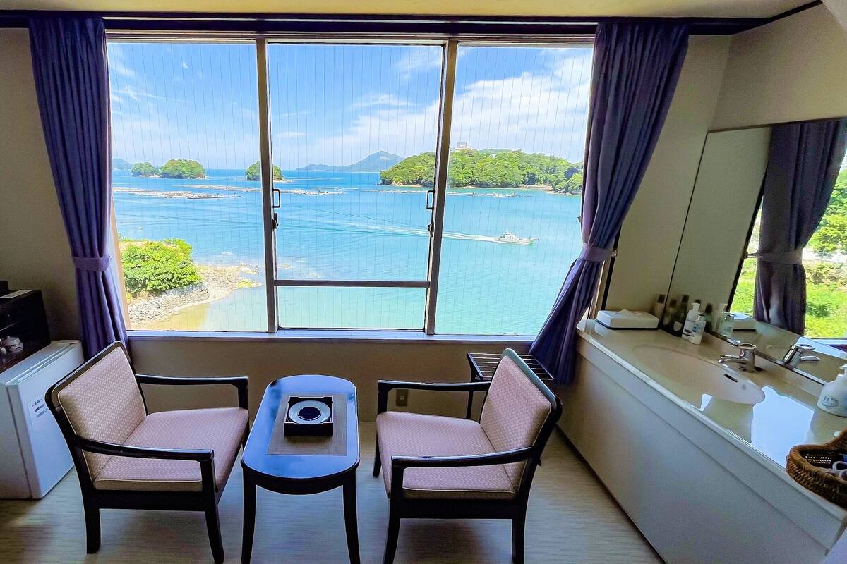 A room with a large window offering a view of a body of water and greenery, two chairs, a small table, and a sink with toiletries.