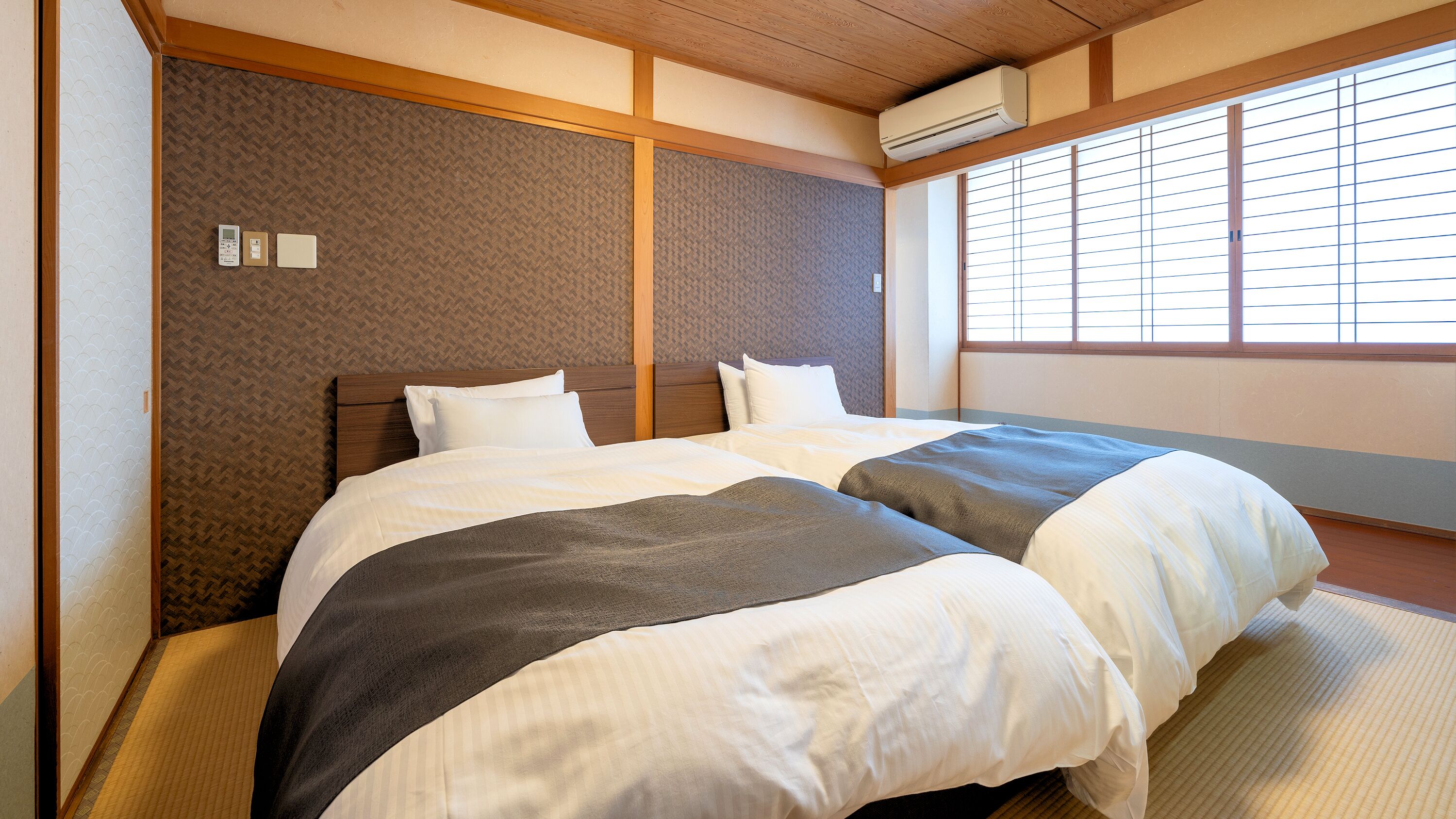 A hotel room with two beds, a wooden headboard, a window with blinds, and an air conditioning unit.