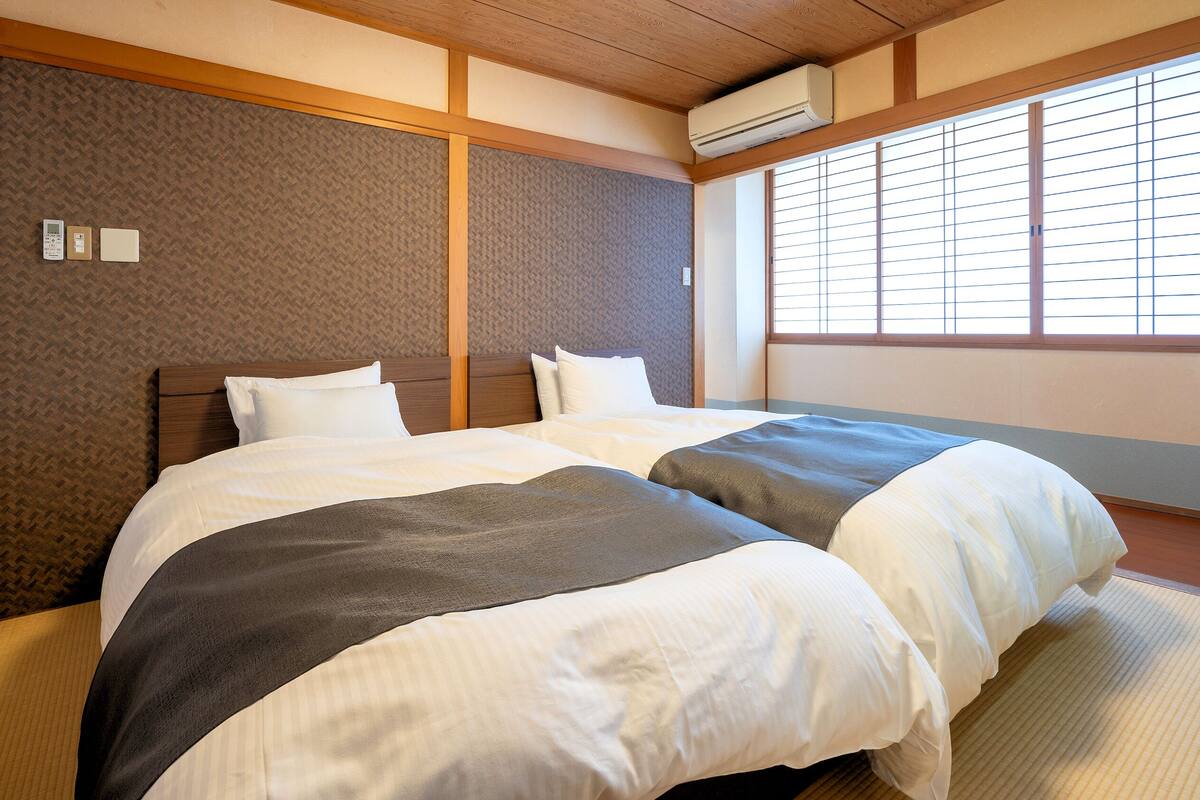 A hotel room with two beds, a wooden headboard, a window with blinds, and an air conditioning unit.