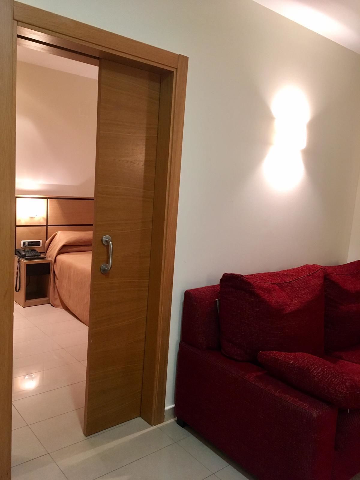 Une pièce avec une porte en bois qui donne sur un espace où se trouve un lit, avec une table de chevet et une lampe. Un canapé rouge est placé contre le mur.