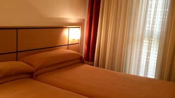 Een hotelkamer met twee bedden, een hoofdeinde met verlichting en een raam met dunne gordijnen.