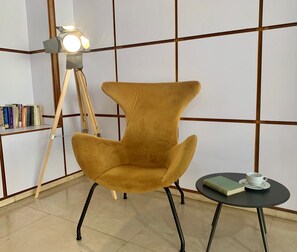 A mustard-colored armchair with black legs, a small round table with a book and a cup, and a floor lamp with a white shade. - Hotel Doña Lola (Castelló de la Plana)