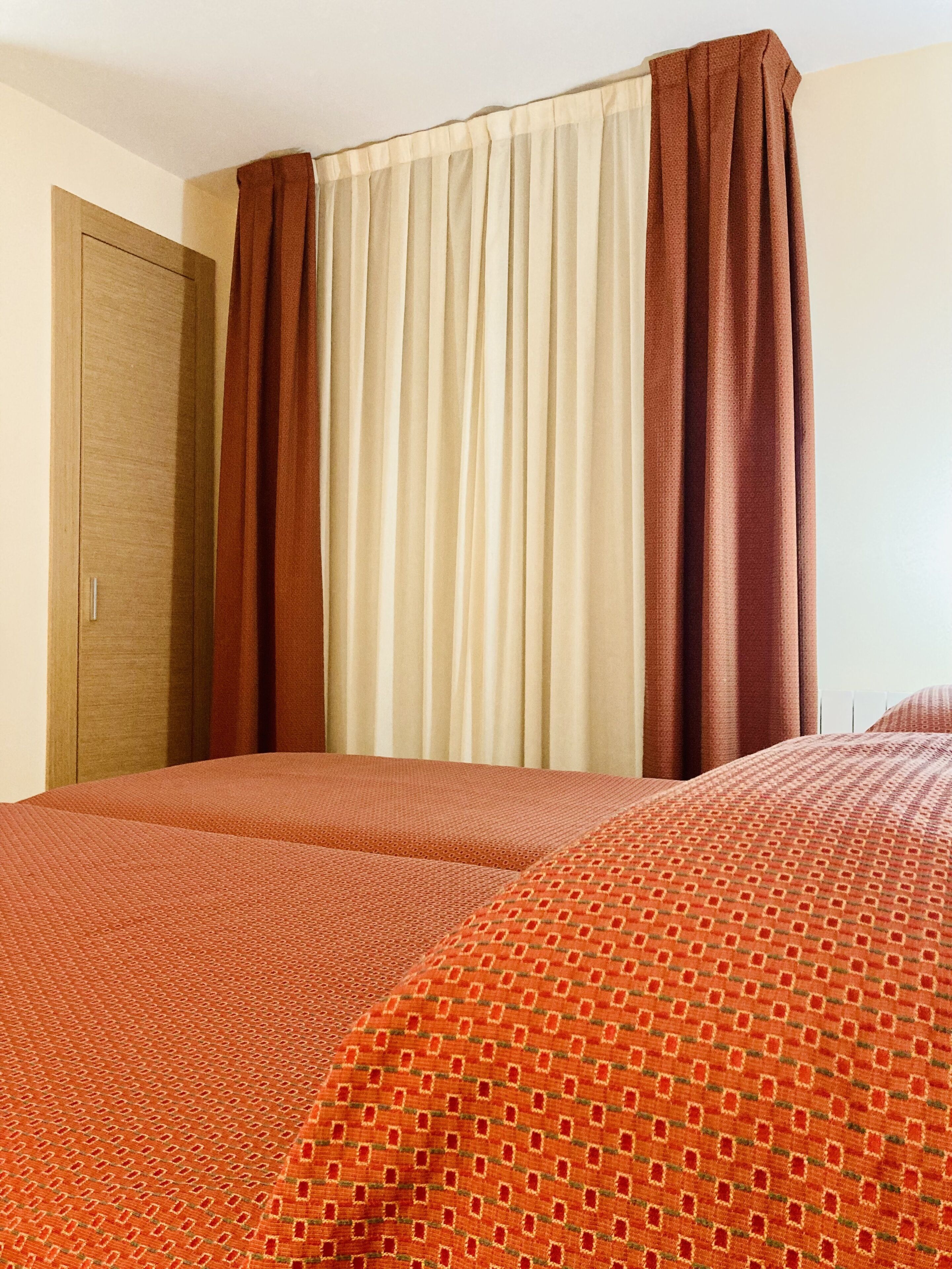 A hotel room with two beds, a wooden door, and a window with beige and brown curtains.