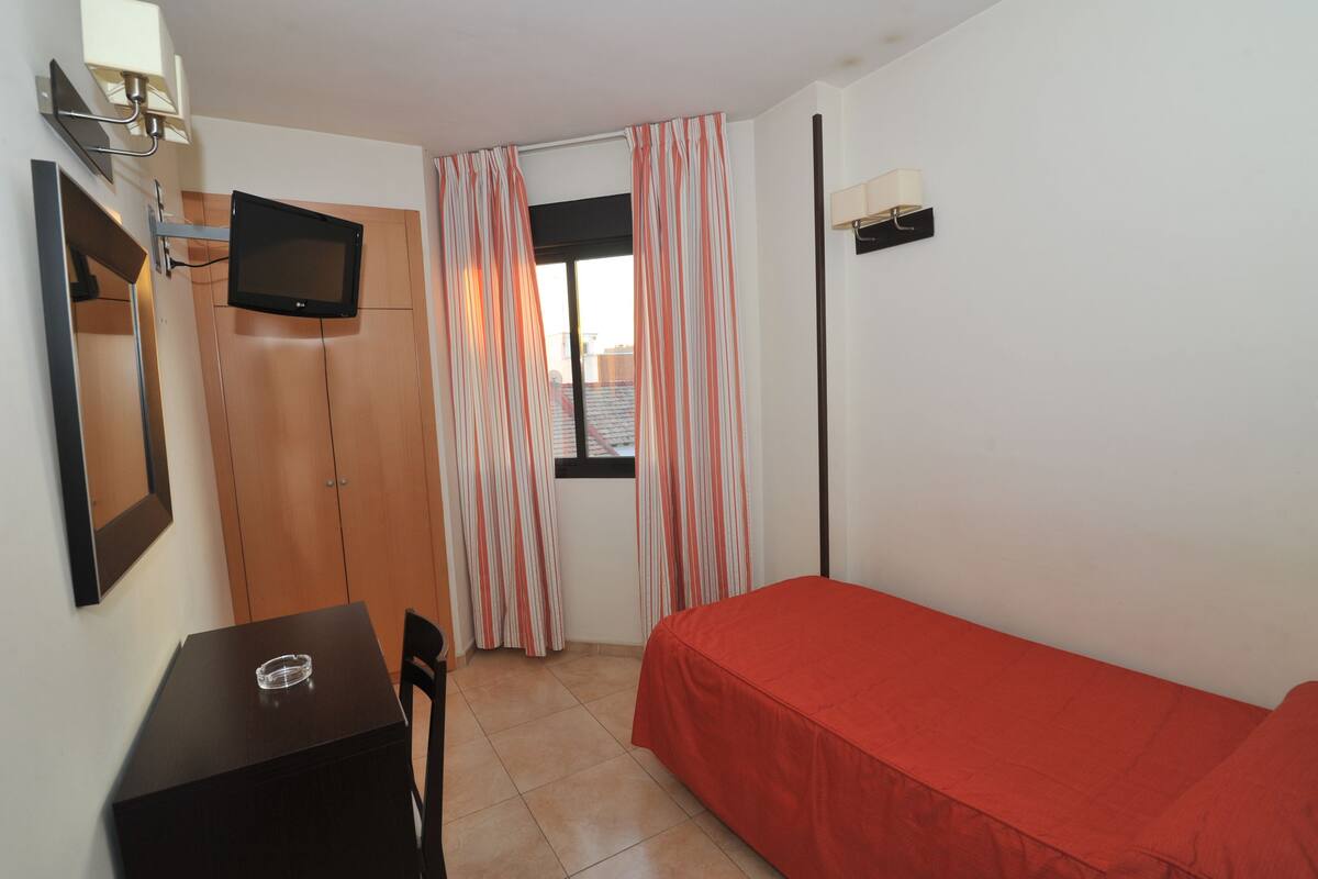 Una habitación pequeña con una cama, un escritorio, una silla, un televisor montado en la pared y un ventanal con cortinas.
