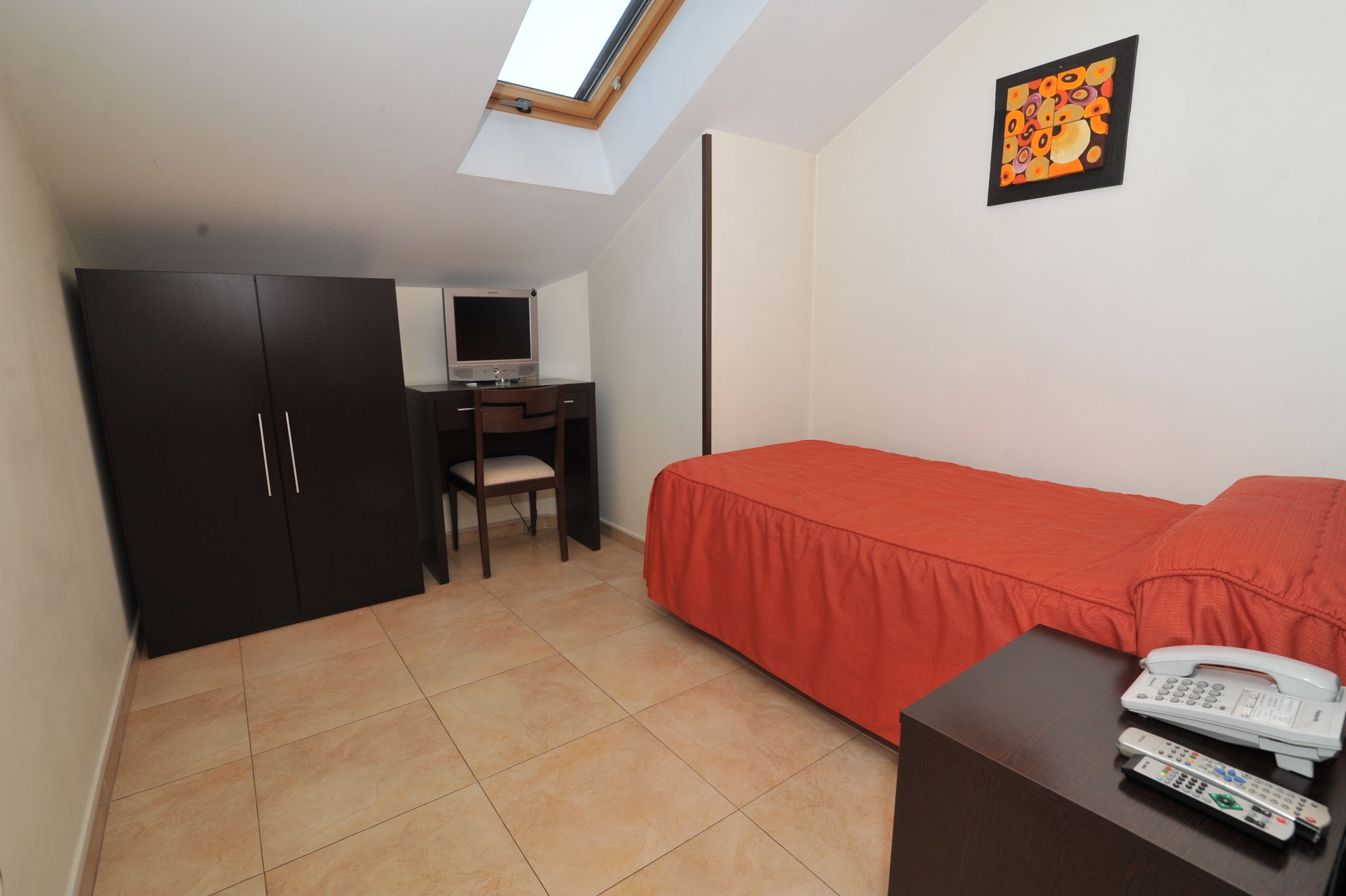 Ein kleines Zimmer mit Bett, Schreibtisch mit Computer, Kleiderschrank und Telefon auf dem Nachttisch.