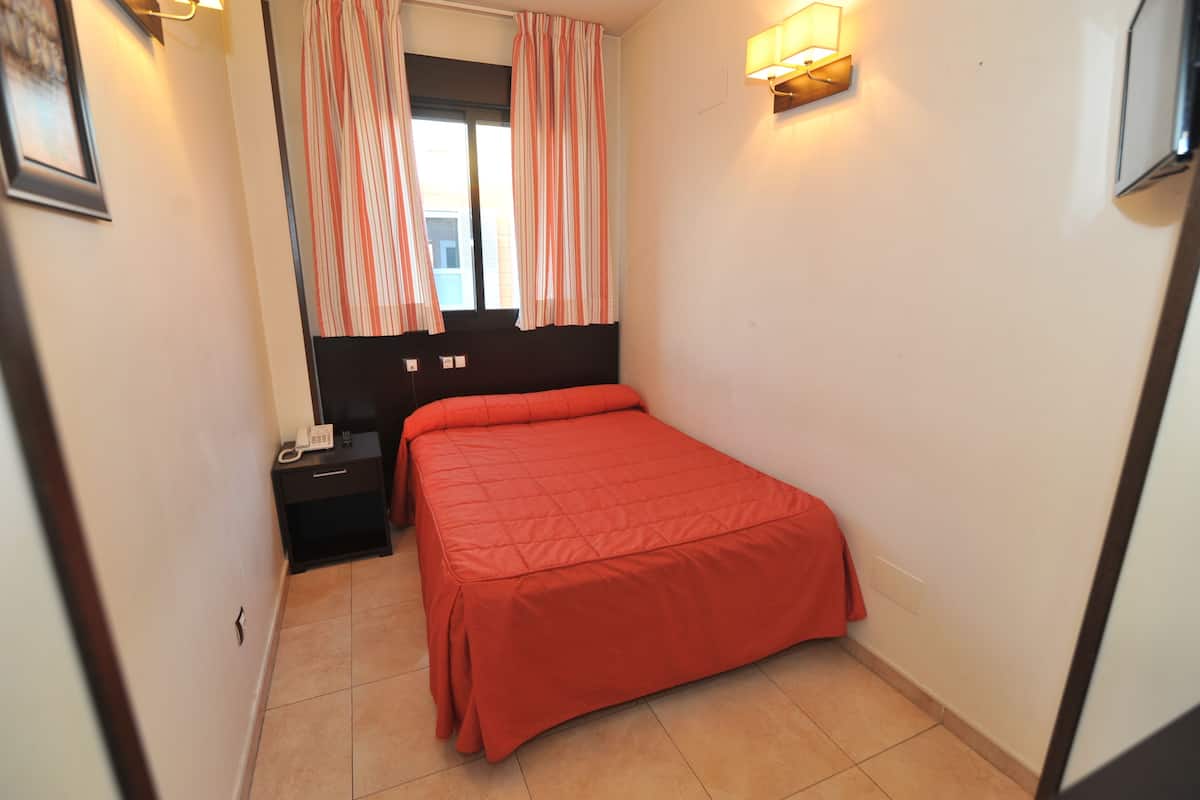 Un dormitorio con cama, mesita de noche, teléfono y ventana con cortinas.