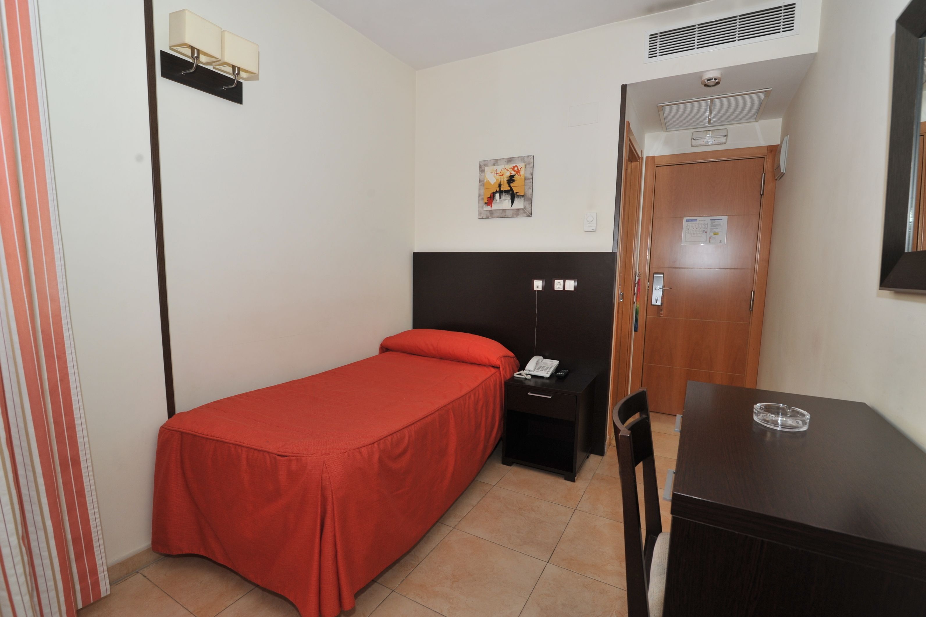 A small, single-bed room with a red bedspread, a wooden nightstand with a phone, a small table with a glass ashtray, and a wooden door with a bulletin board.