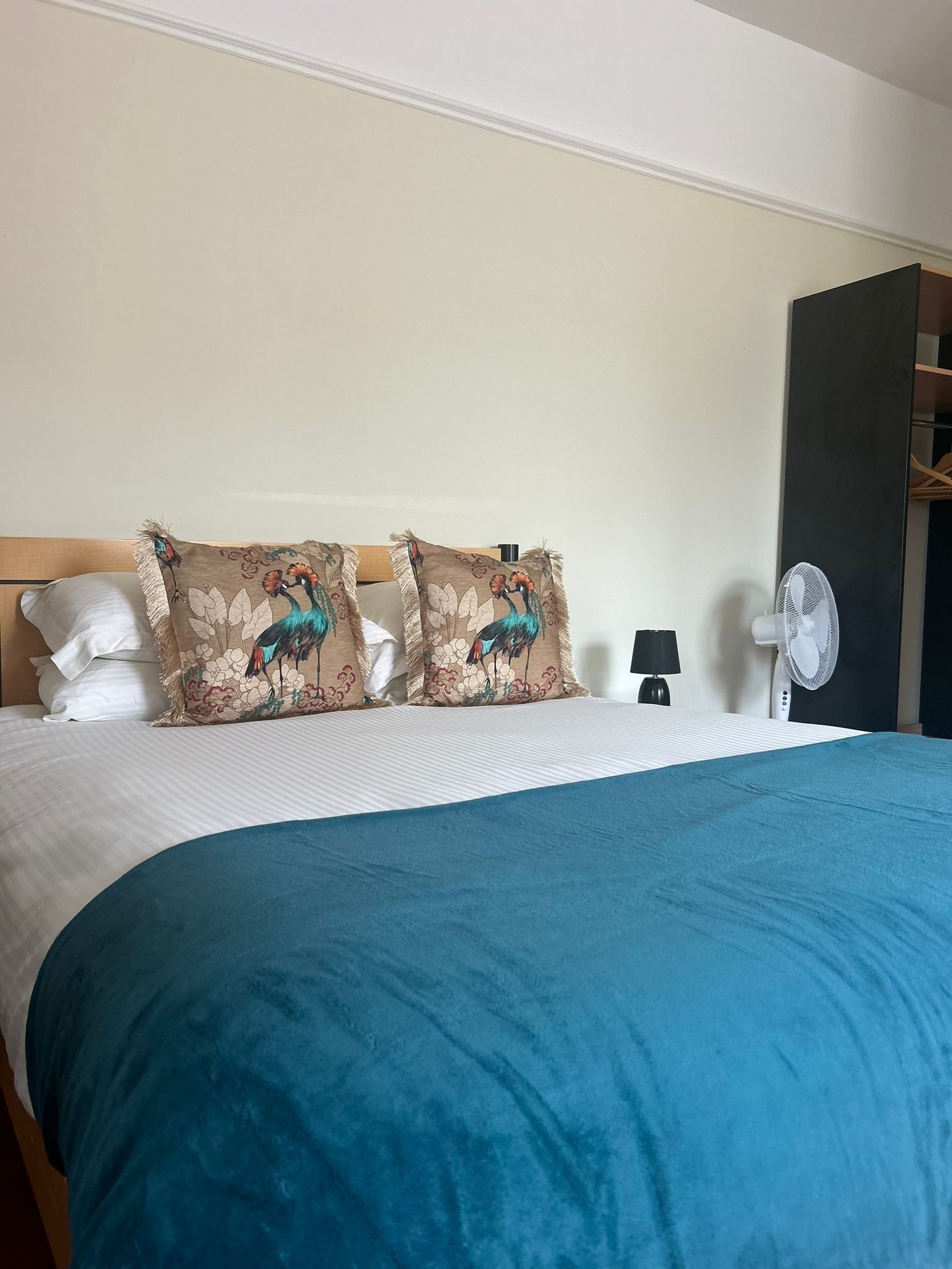A hotel room with a bed, two decorative pillows with peacock designs, a blue bedspread, a wall-mounted fan, and a nightstand with a lamp.